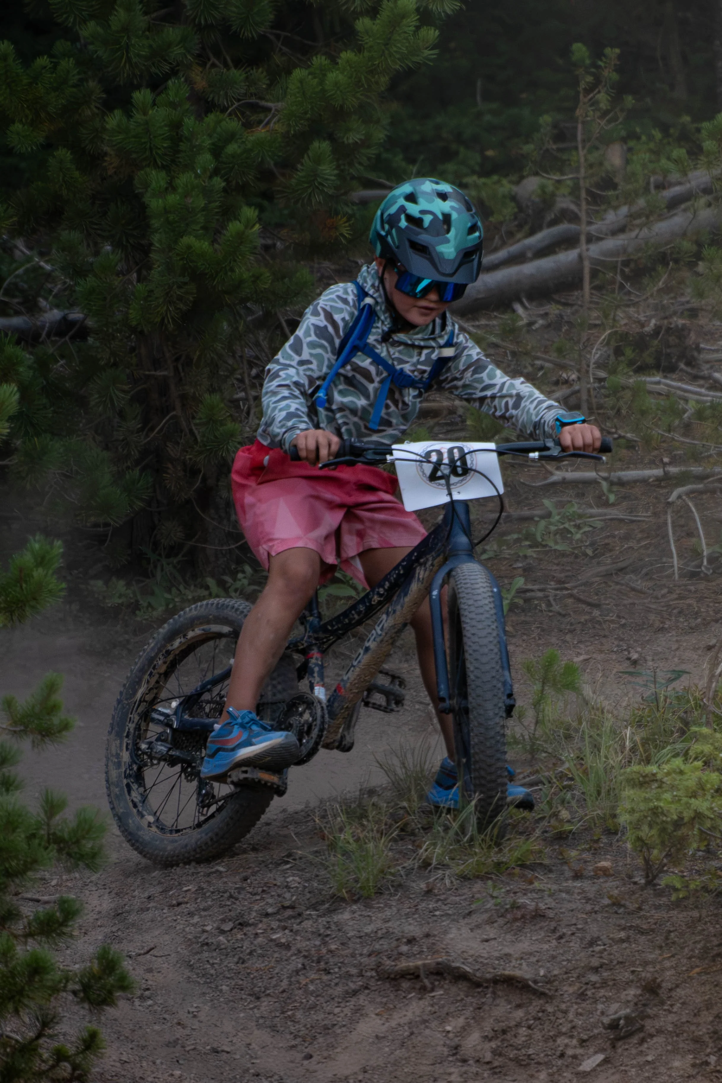 A young boy wearing a blue helmet, blue sunglasses, a camouflage-patterned jacket, and pink shorts, riding a mountain bike on a dirt trail through a forest with green trees.