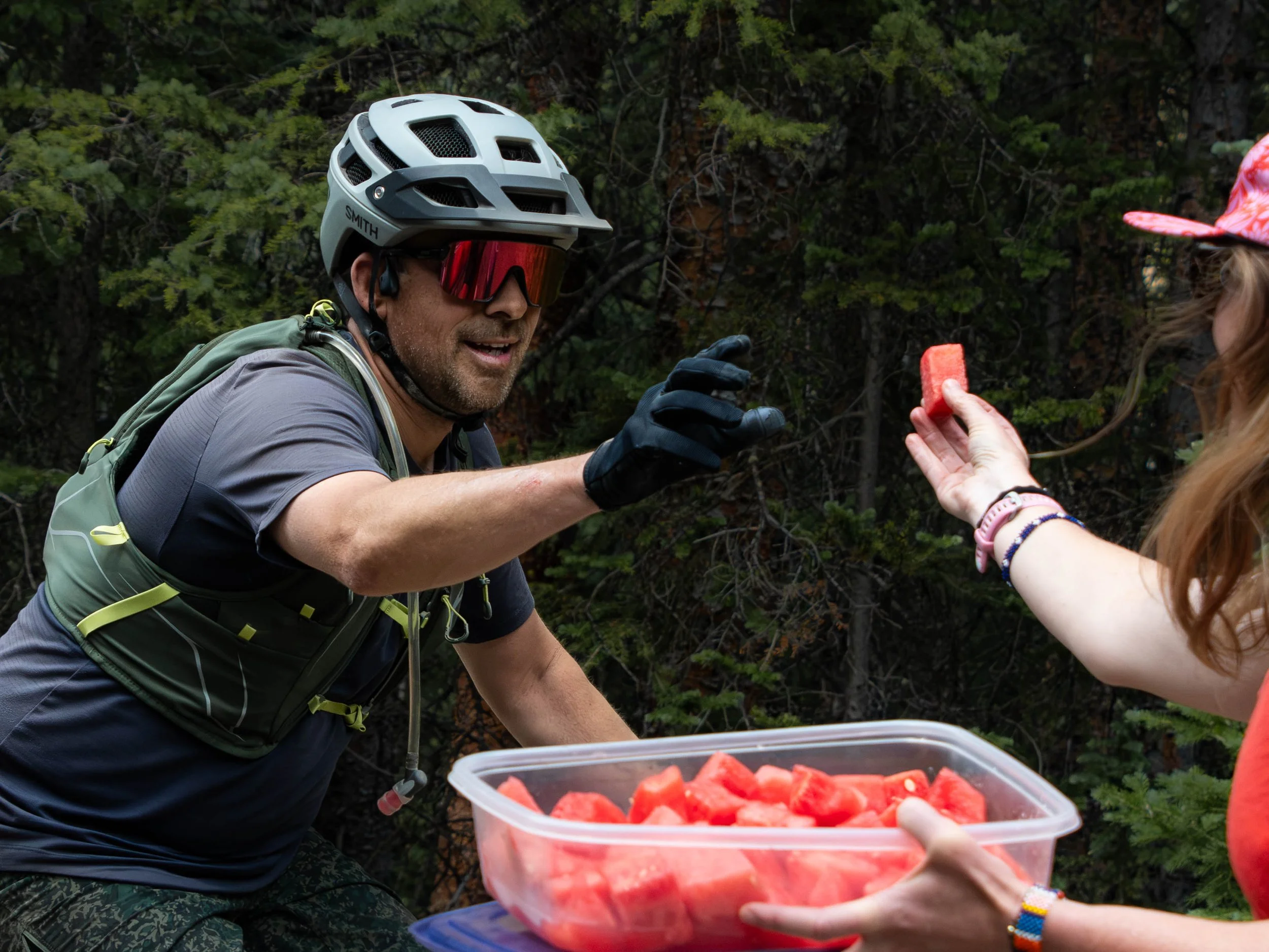 A man wearing a bike helmet with the brand 'SMITH' and red riding goggles is reaching out to receive a slice of watermelon from a woman. The woman’s hand is visible holding the watermelon, and she is wearing a pink watch and colorful bracelets. The b