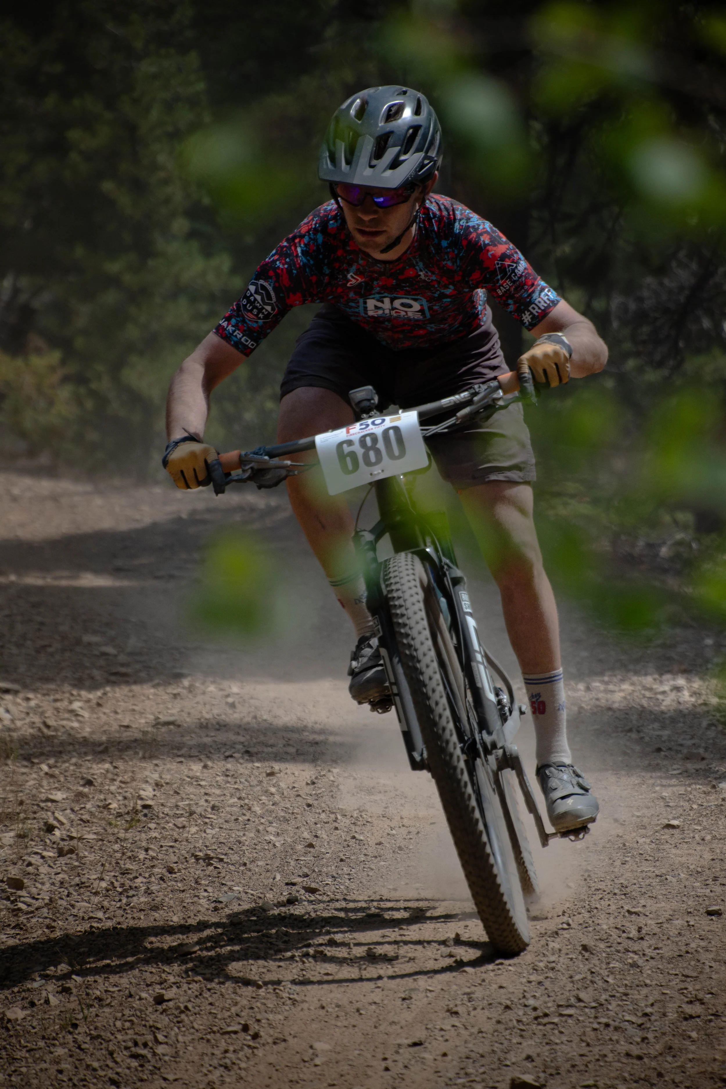 A person riding a mountain bike on a dirt trail in a wooded area, wearing a helmet, sunglasses, a colorful jersey, and gloves, with a race number "680" attached to the bike.
