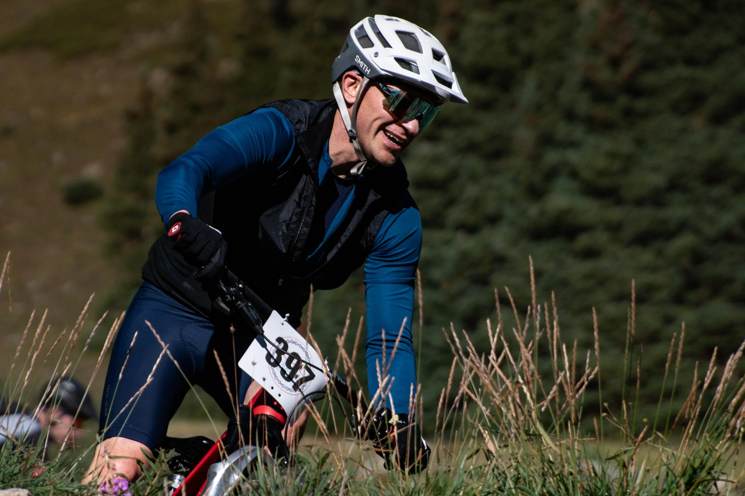Male mountain biker wearing a white helmet with black vents, reflective sunglasses, and a blue and black biking outfit, riding through grassy terrain with tall grass in the foreground and a blurred green forest background.