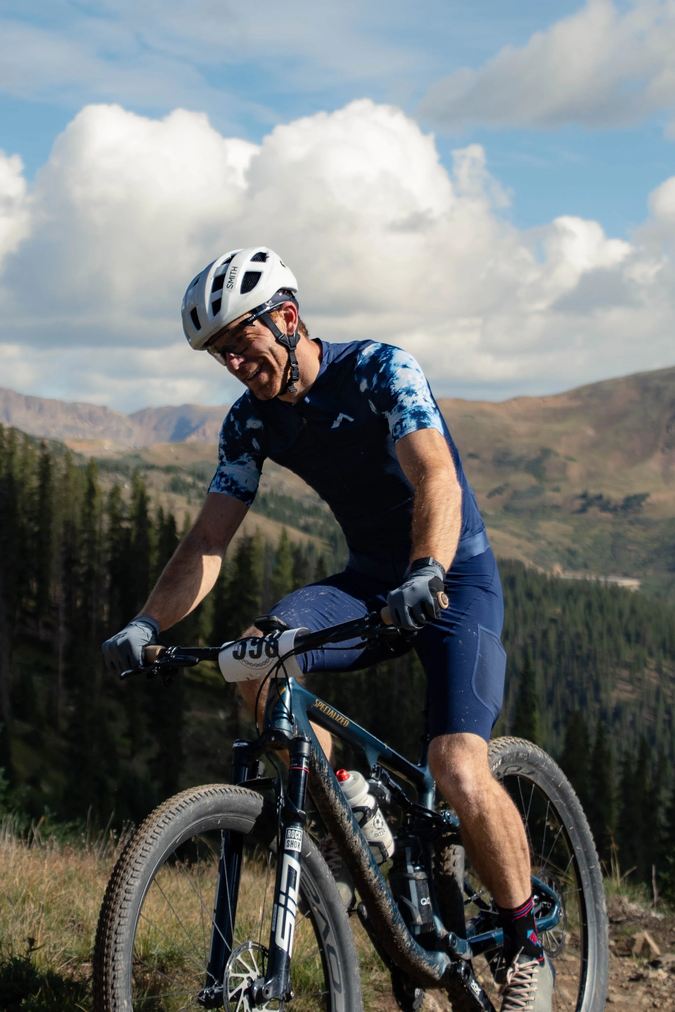 A man riding a mountain bike outdoors in a mountainous area with pine trees and clouds in the sky.