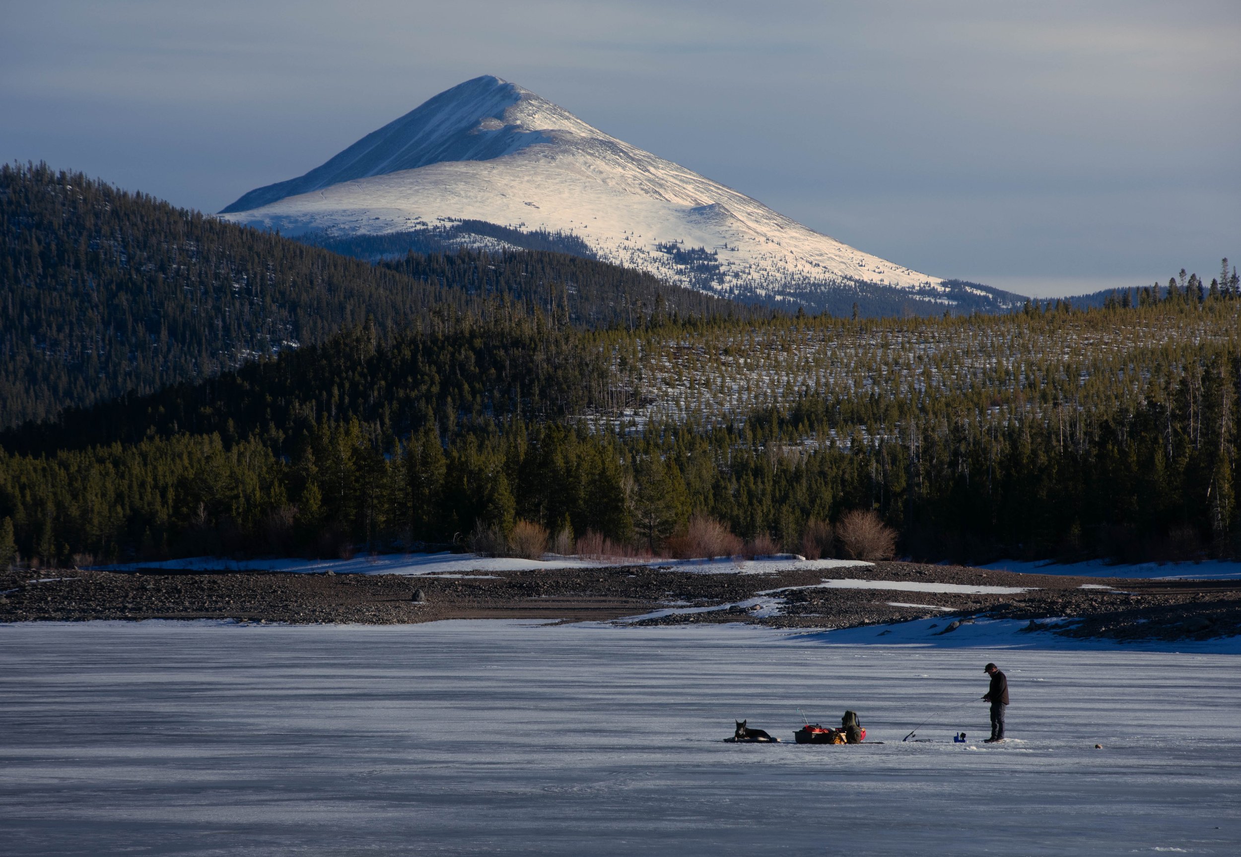A snow-covered mountain and forest in the background with a frozen body of water in the foreground. Two people and a dog are on the ice, engaging in winter activities.