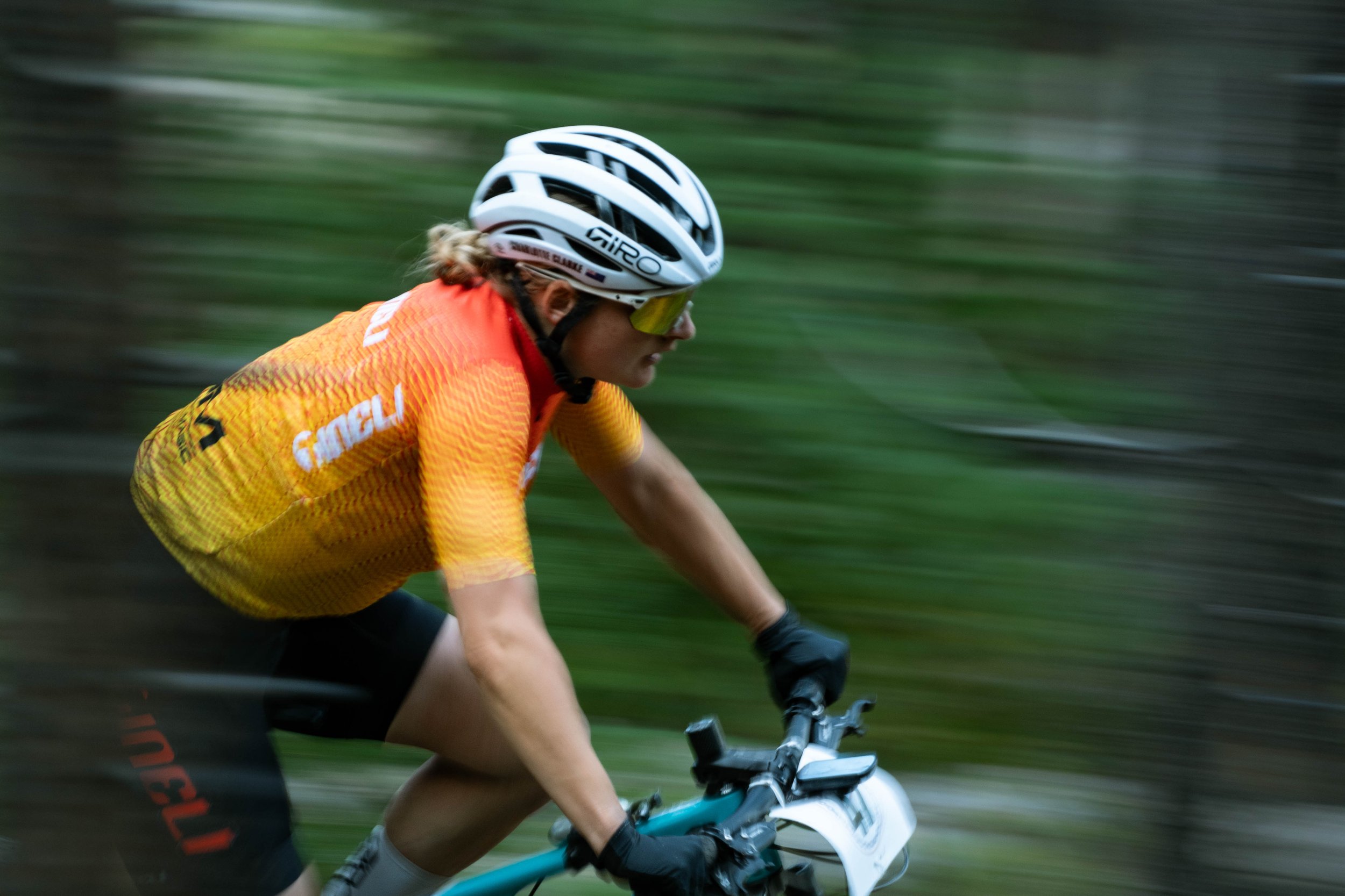 A cyclist wearing an orange jersey and a white helmet riding a mountain bike through a forest trail.