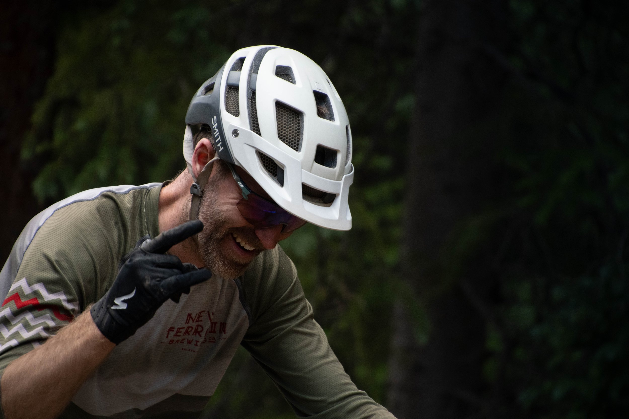 A man wearing a white helmet with black and gray accents, sunglasses, a beige cycling jersey with red and white zigzag pattern on the sleeve, and black gloves, is smiling and gesturing with his right hand.
