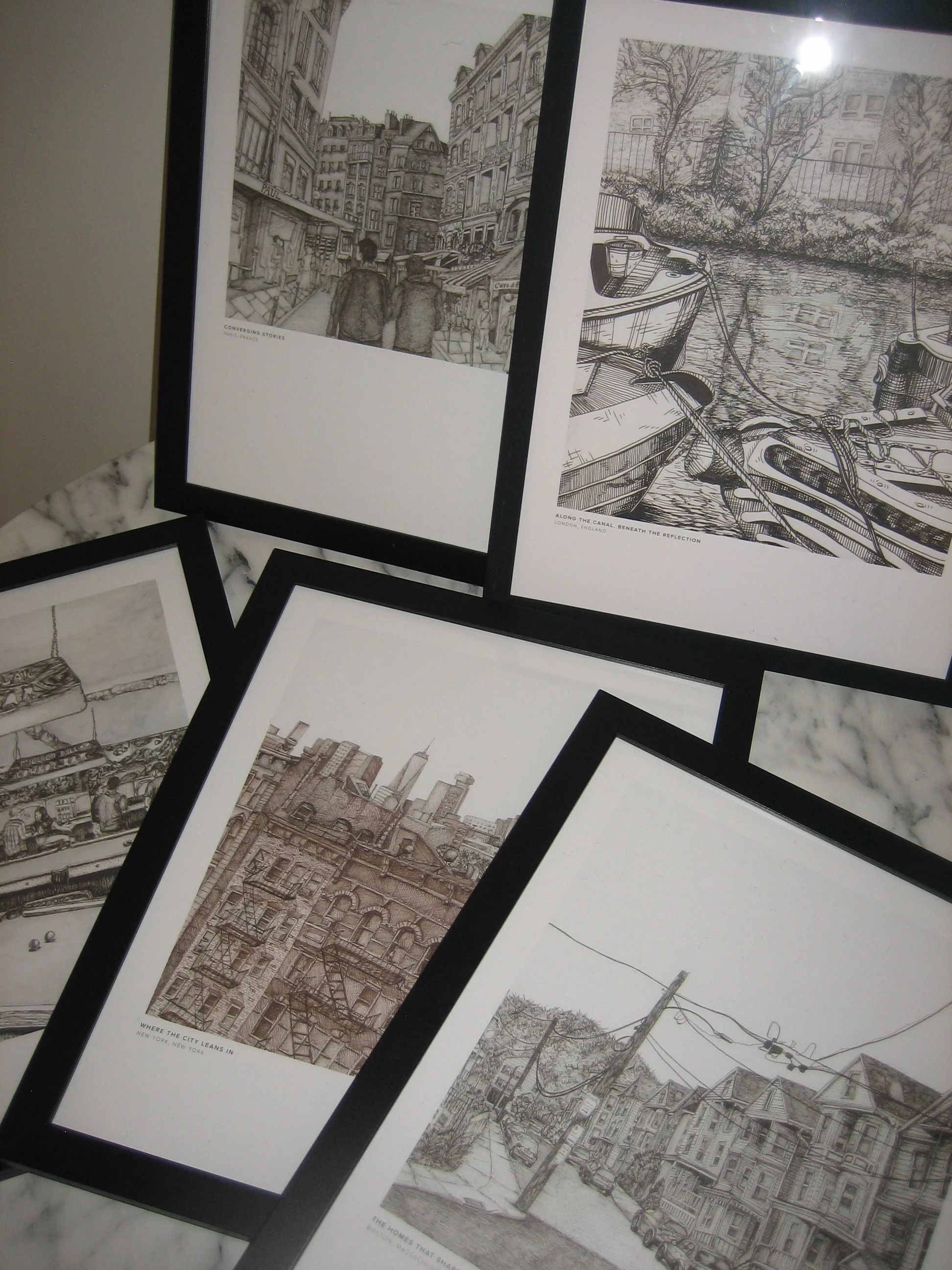 Collection of framed black and white line drawings of cityscapes, including a canal scene with boats, a busy street with buildings, and an industrial area with power lines.