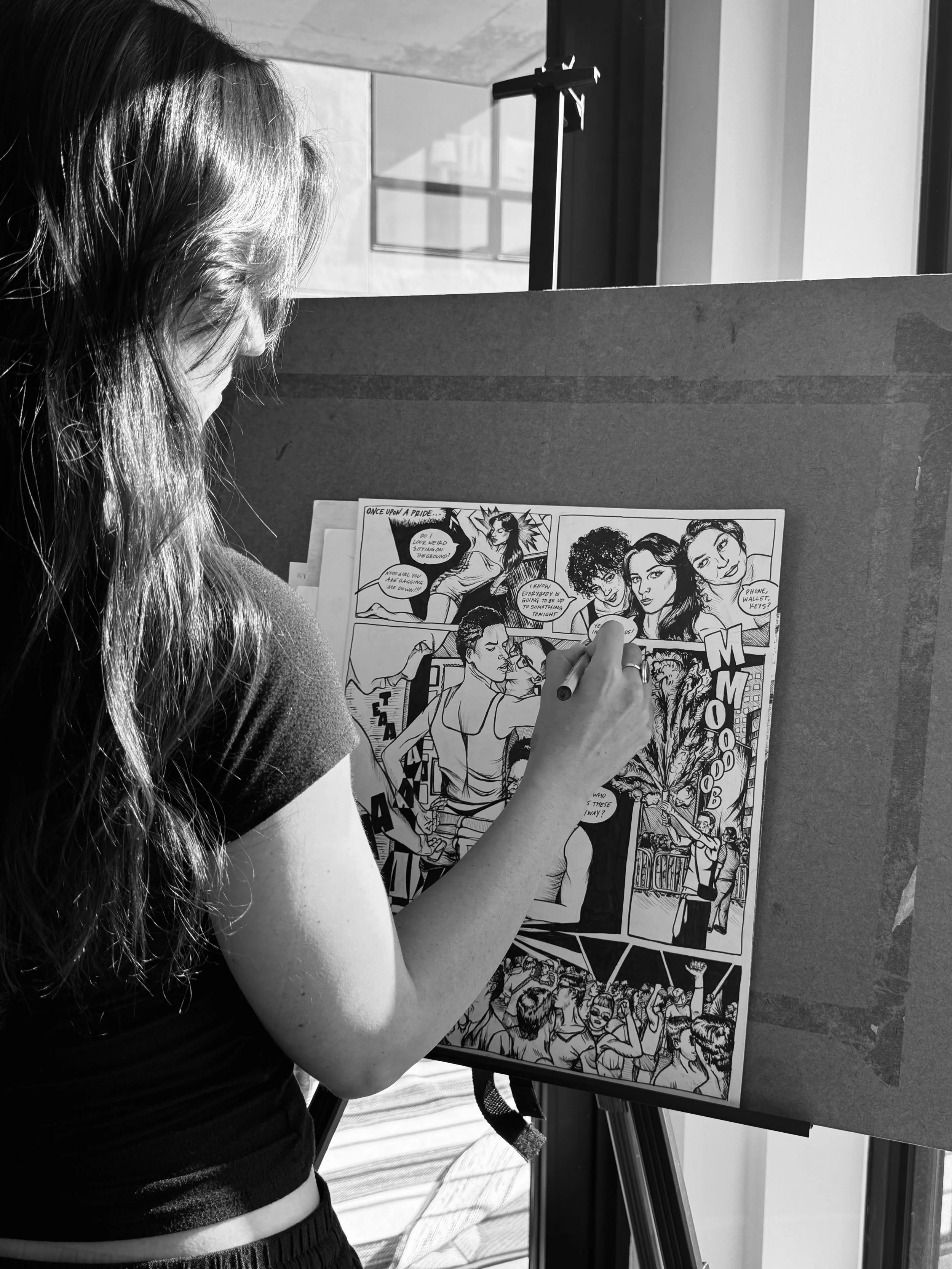 A person with long hair is drawing on a large comic book page on an easel, with a window in the background.