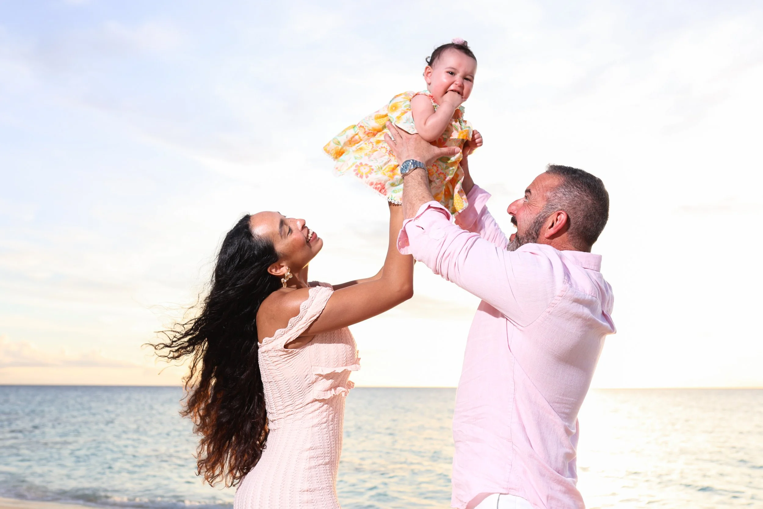 A family of three enjoying a moment together on the beach during sunset. The father is lifting the toddler girl into the air, the mother is smiling and holding her from below, all facing each other with the ocean and sky in the background.