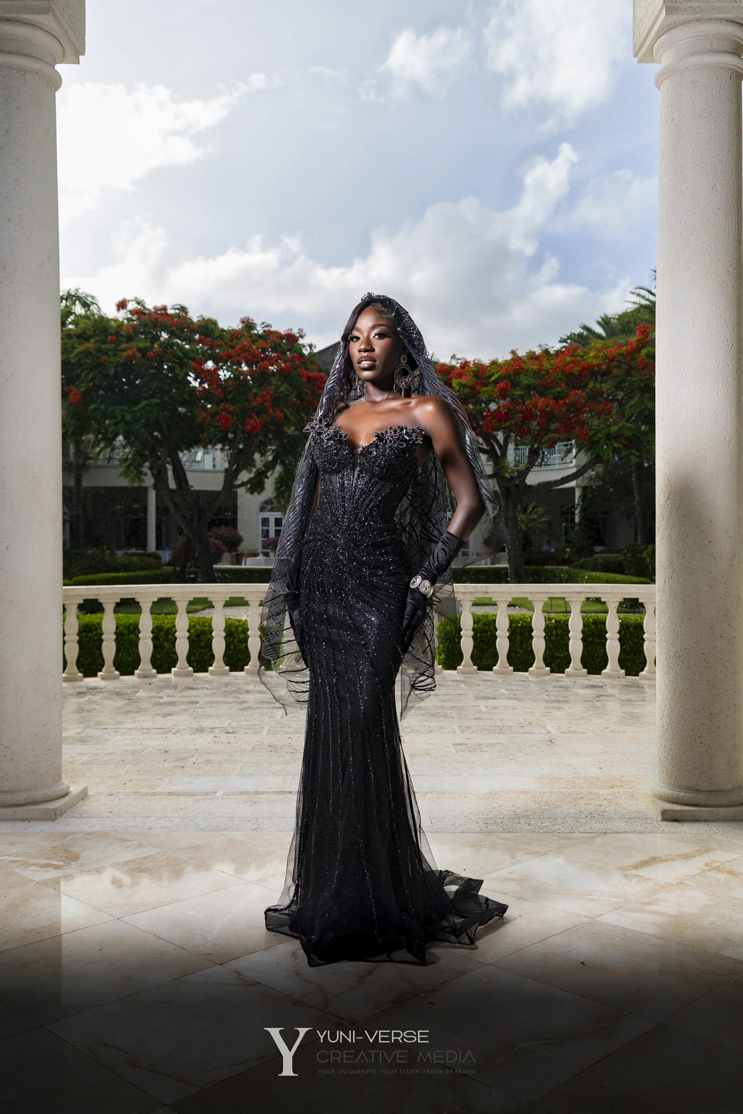 A woman dressed in a black, strapless, sequined evening gown stands under white columns with a garden and trees with red flowers behind her. The sky is partly cloudy, and she is wearing earrings, gloves, and a wristwatch.