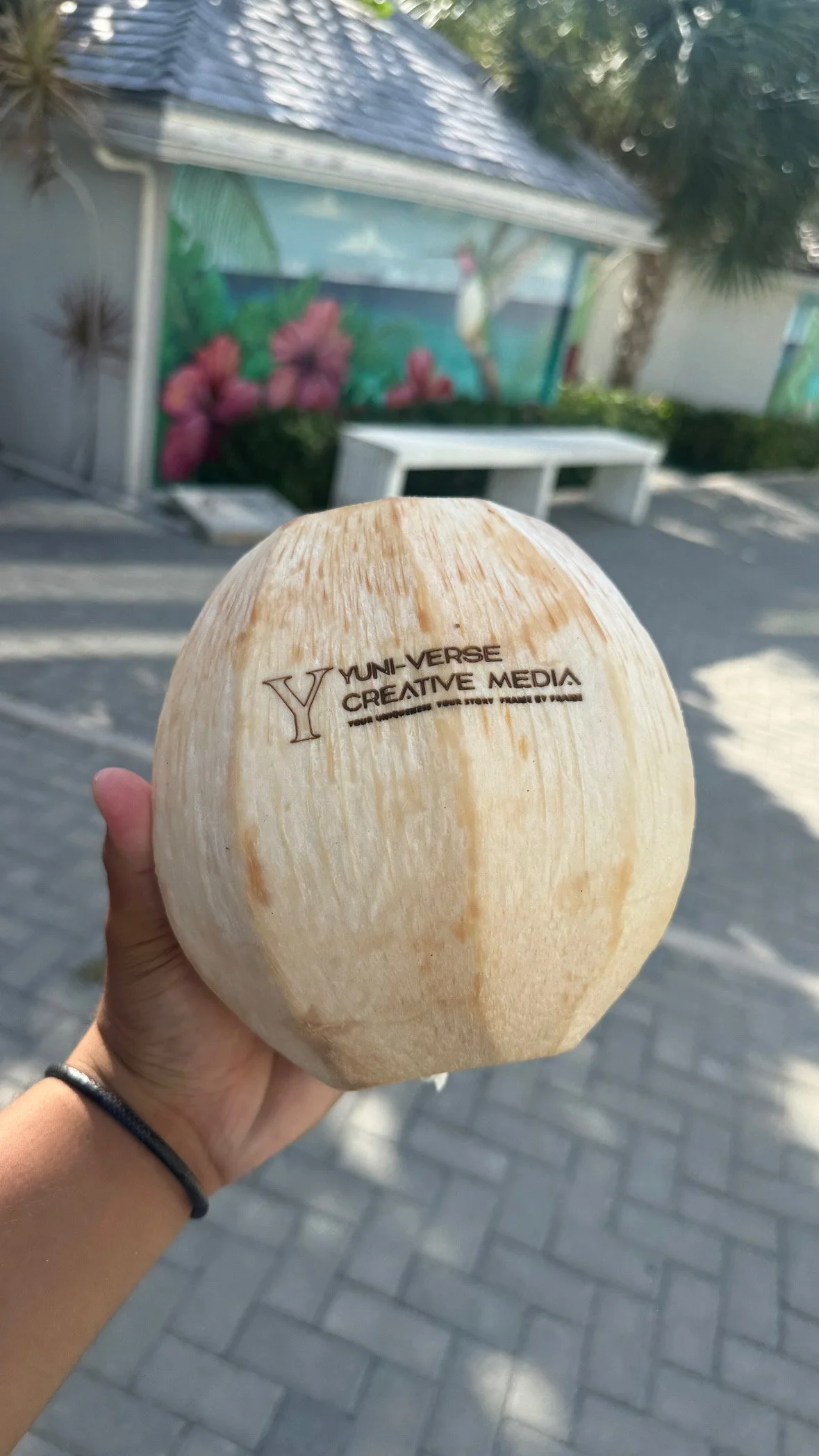 A person holding a large coconut with the words 'YUNI-VVERSE CREATIVE MEDIA' printed on it. In the background, there is a mural with pink flowers and green foliage, and some trees and a bench are visible.