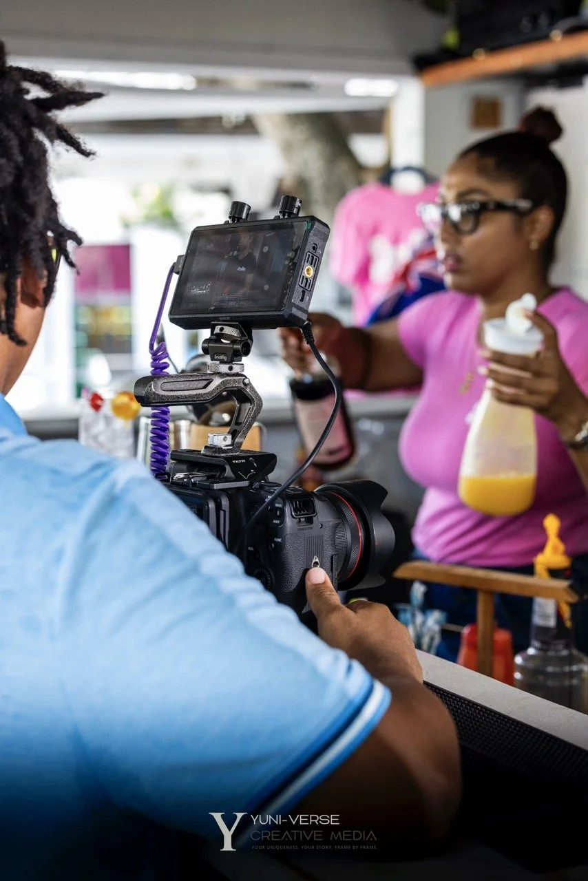A person filming a woman holding a drink and a spray bottle with yellow liquid inside. The woman is wearing a pink shirt and glasses, and the scene appears to be in a casual setting, possibly a food truck or outdoor stand.