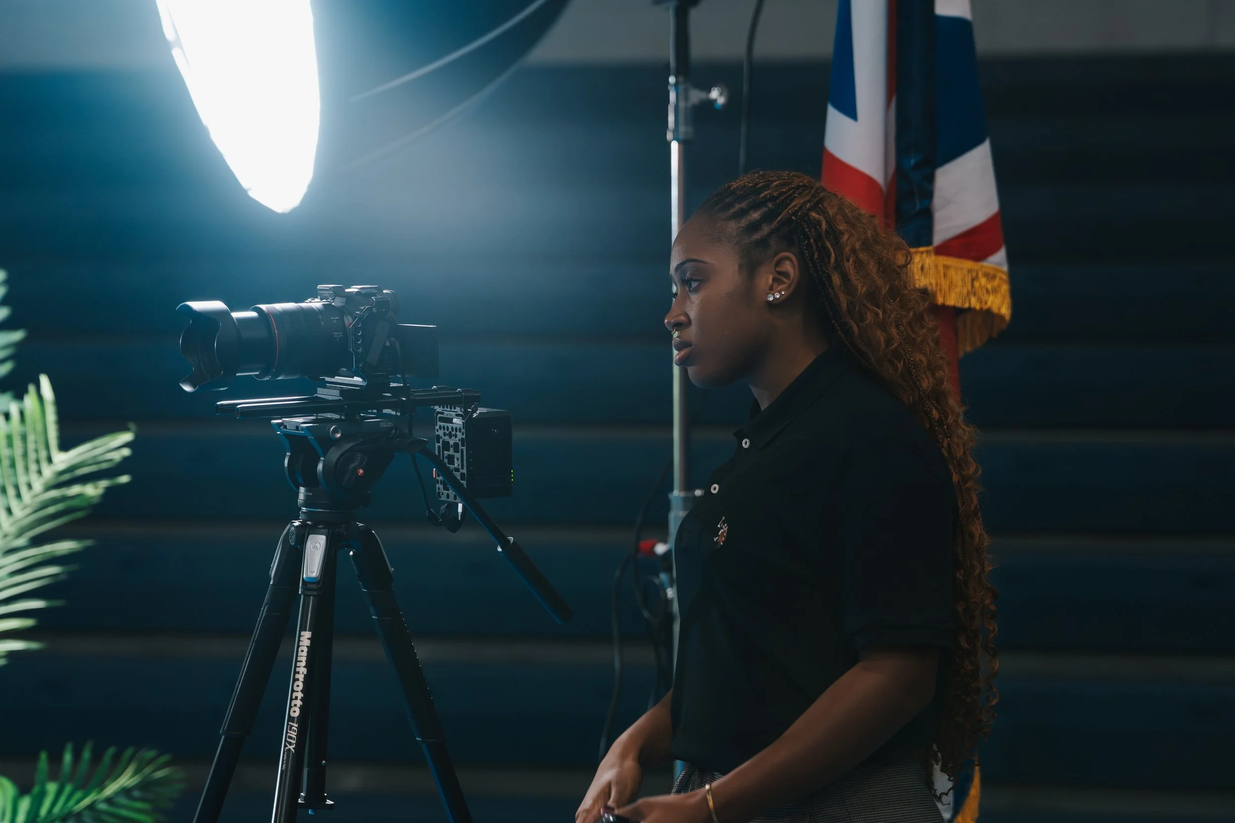 A woman with long, curly hair filming with a professional camera mounted on a tripod inside a studio. There is a bright light source to the left and a Union Jack flag in the background.