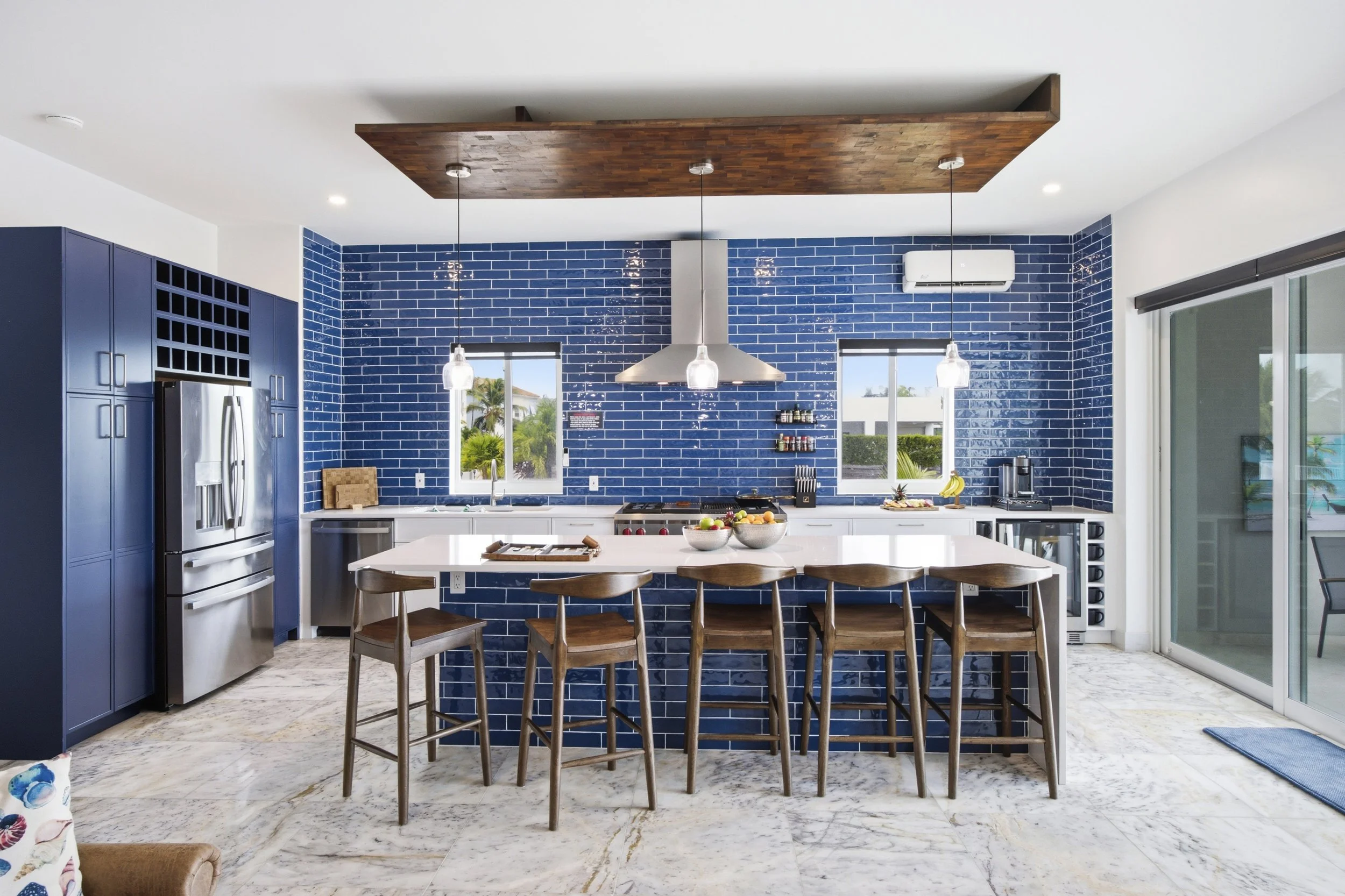 Modern kitchen with blue tiled walls, white countertops, a central island with bar stools, large windows, and stainless steel appliances.
