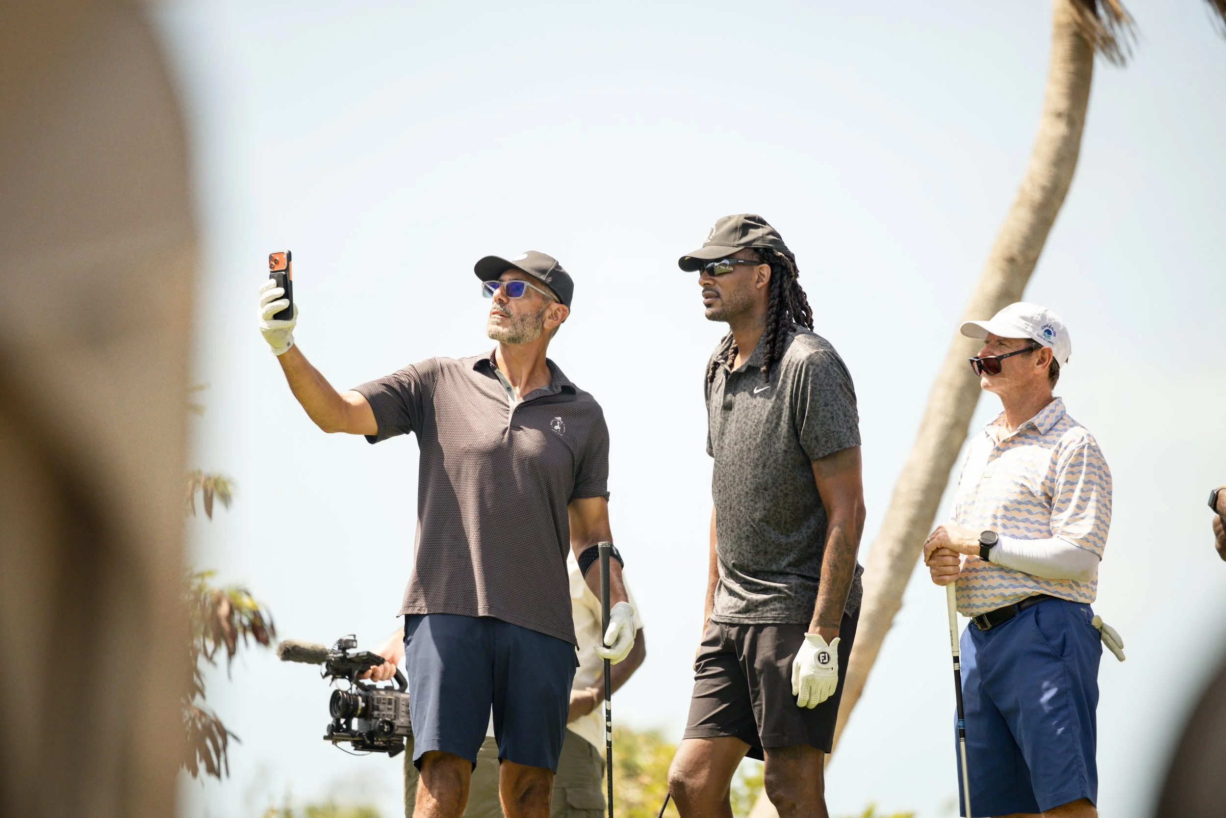 Three men standing on a golf course. One man is taking a selfie, another man is looking into the distance, and the third man is holding a golf club, all dressed in golf attire with hats and sunglasses. The photo is taken outdoors in bright sunlight.