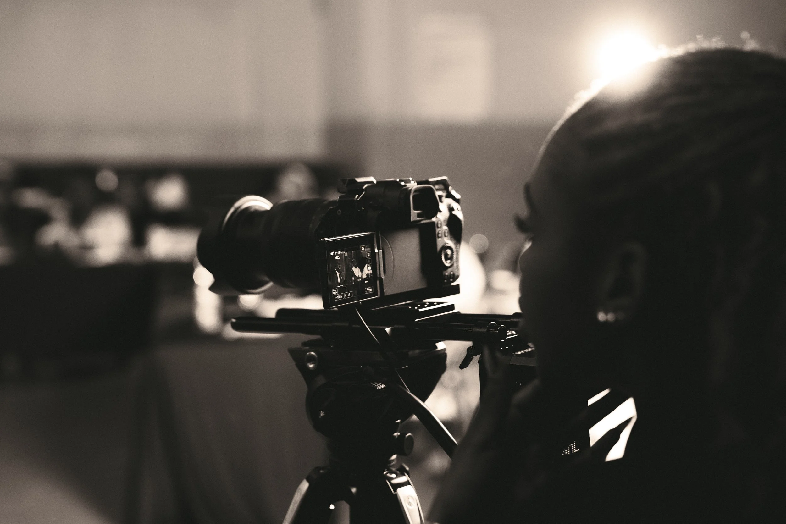 A person operating a professional camera mounted on a tripod, with a blurred background and a bright light source behind their head.