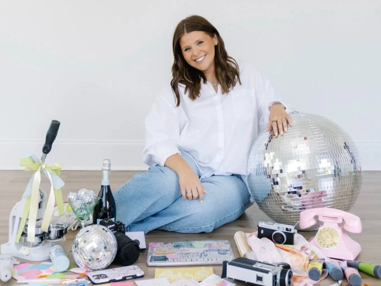 A woman sitting on the floor, smiling, with a large disco ball, cameras, a vintage pink rotary phone, wine bottles, and various party accessories surrounding her.