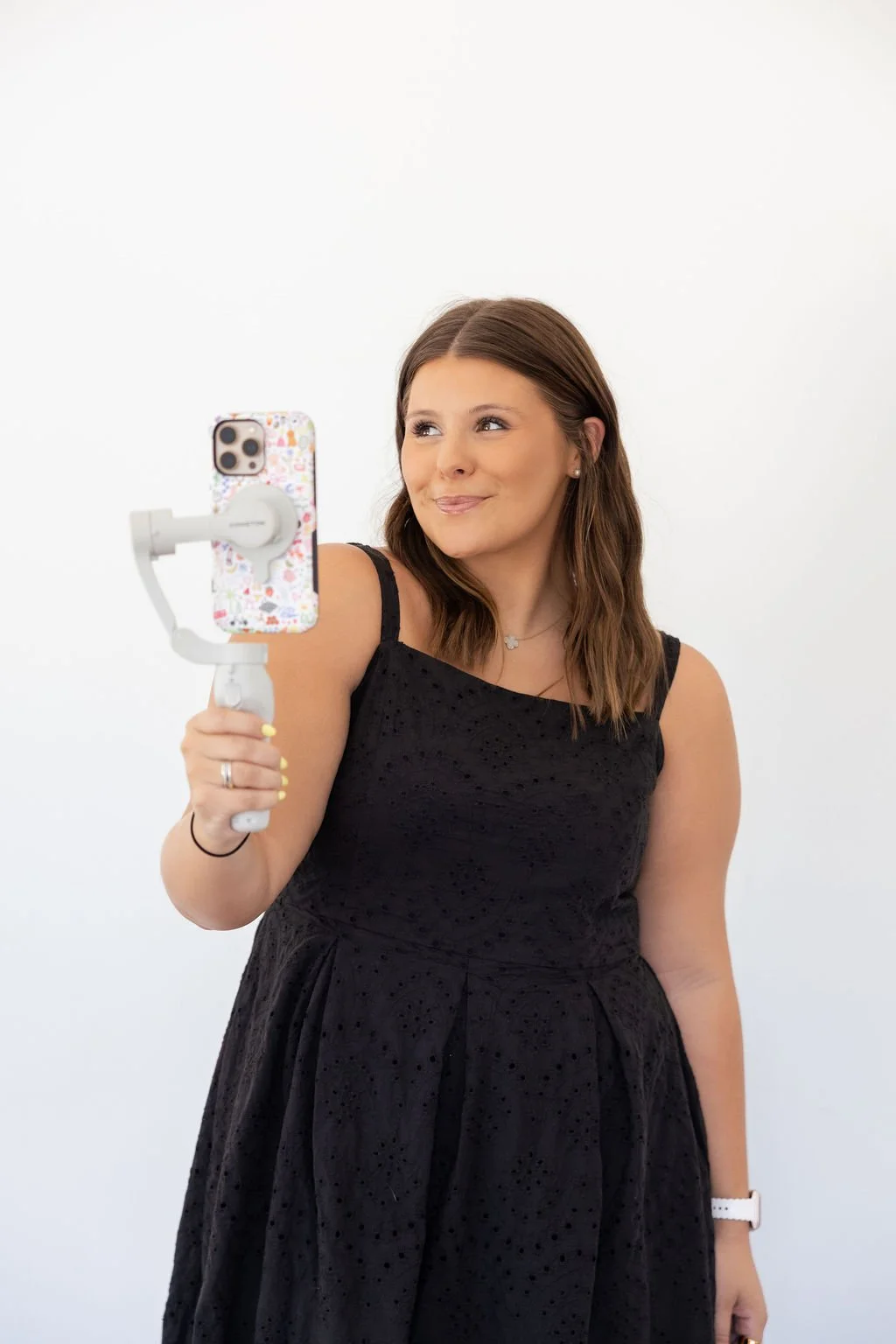 A young woman with brown hair, wearing a black dress, takes a selfie with her smartphone mounted on a white gimbal against a plain white background.