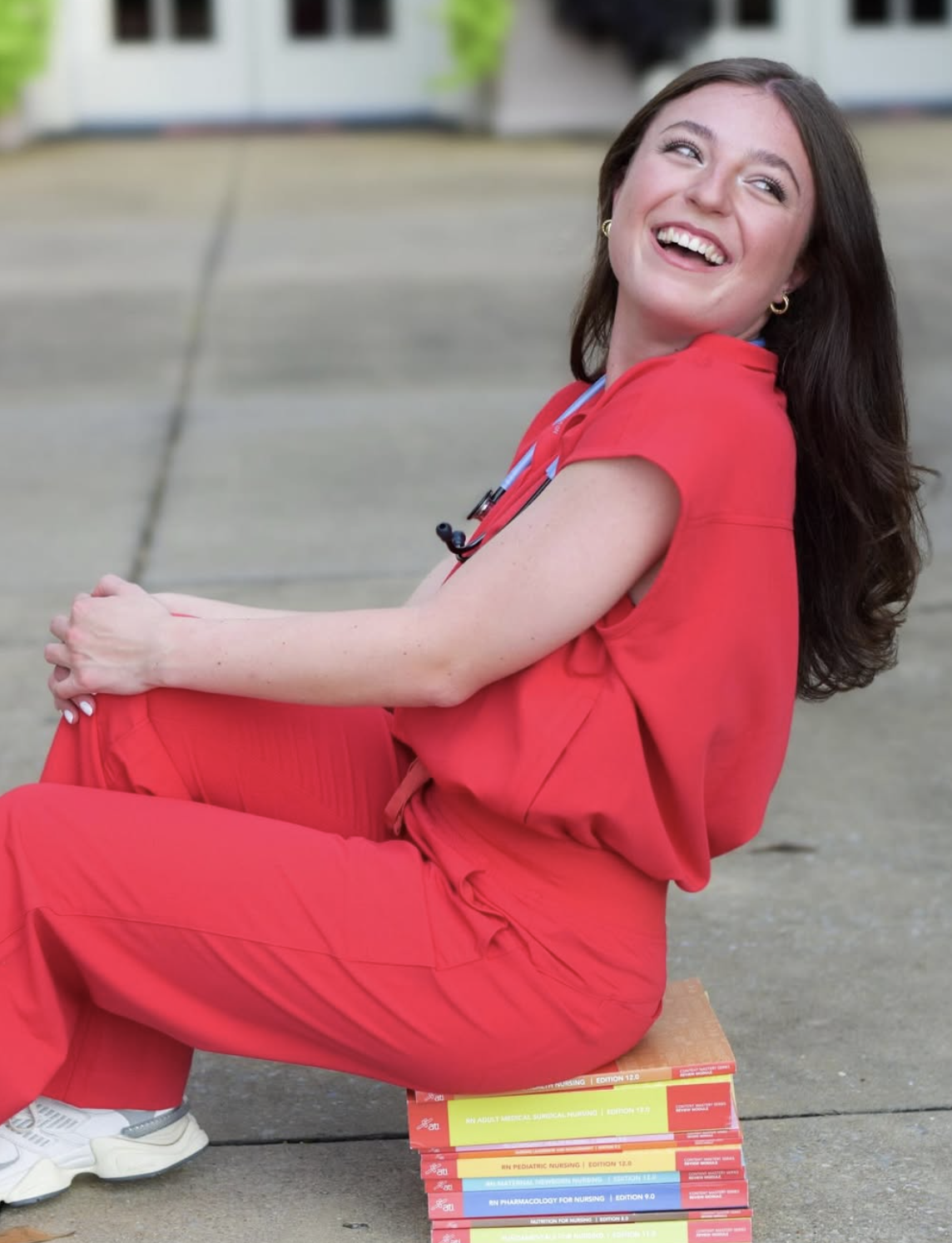 A smiling woman in red nursing scrubs sitting on a stack of nursing textbooks outdoors.