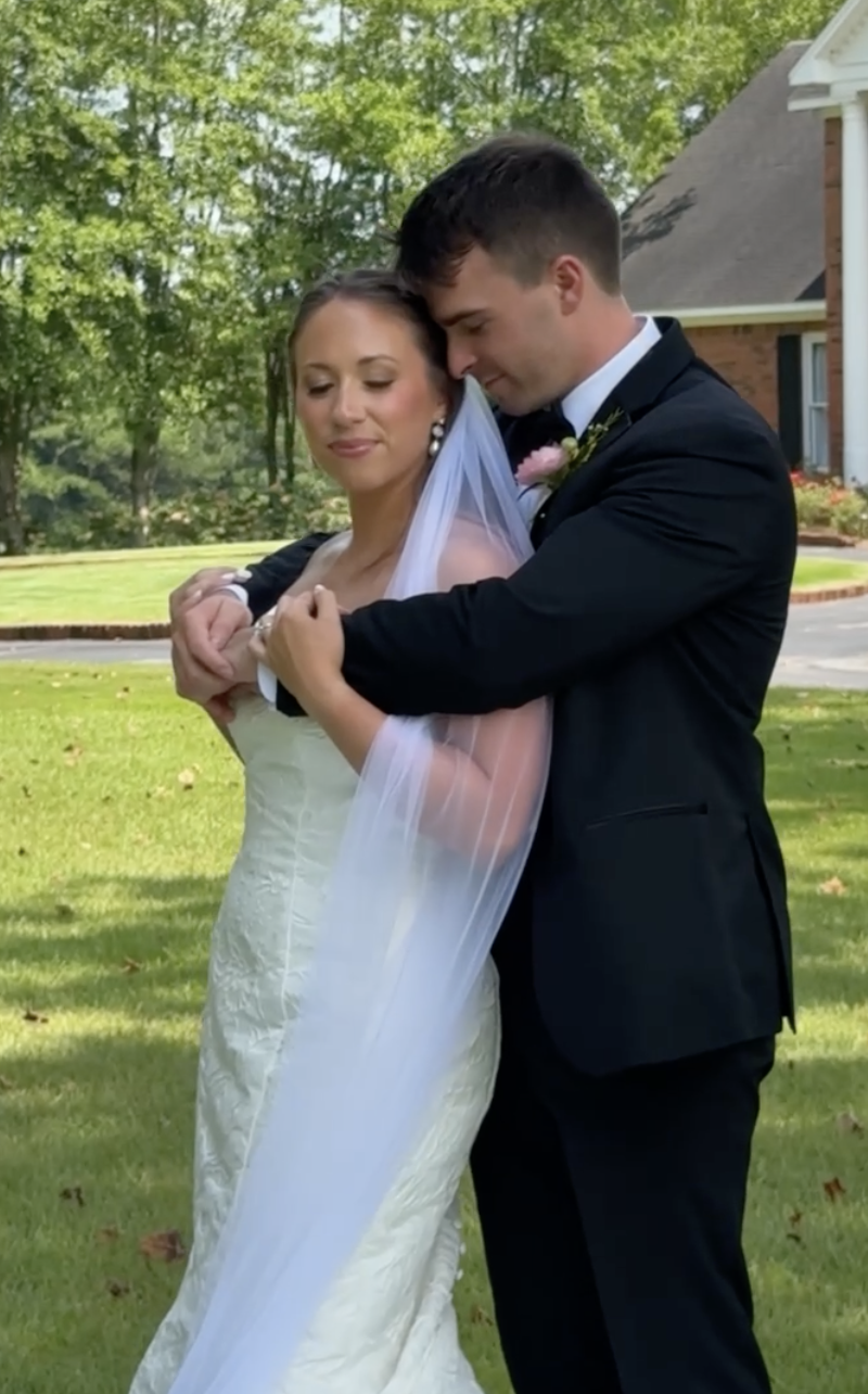 A bride and groom embrace outdoors on their wedding day, with the groom gently holding the bride as they look content and serene, in front of a green lawn and a house in the background.