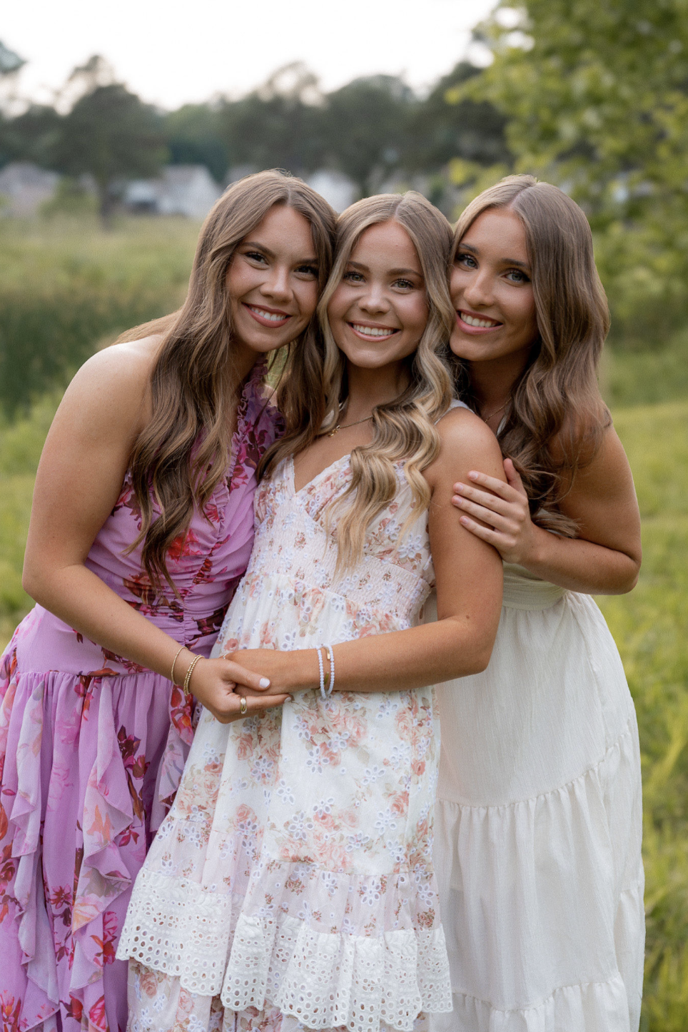 Three young women in summer dresses smiling and hugging each other outdoors in a grassy field with trees and houses in the background.