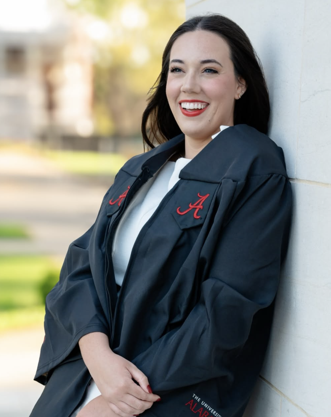 A smiling woman in a graduation gown with an Alabama 'A' emblem, leaning against a wall outdoors.