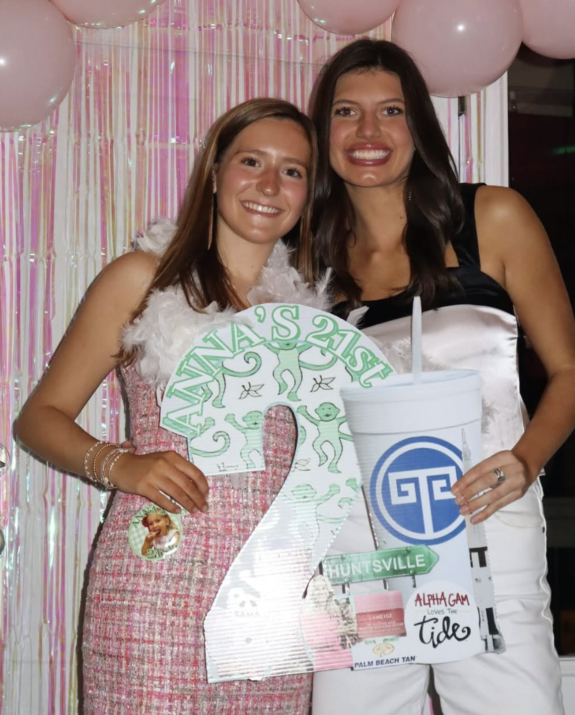 Two young women smiling at a celebration, holding a large birthday sign that says 'Ariana's 21st' with angel illustrations and a photo of a young girl, in front of pink balloons and shiny pink streamers.