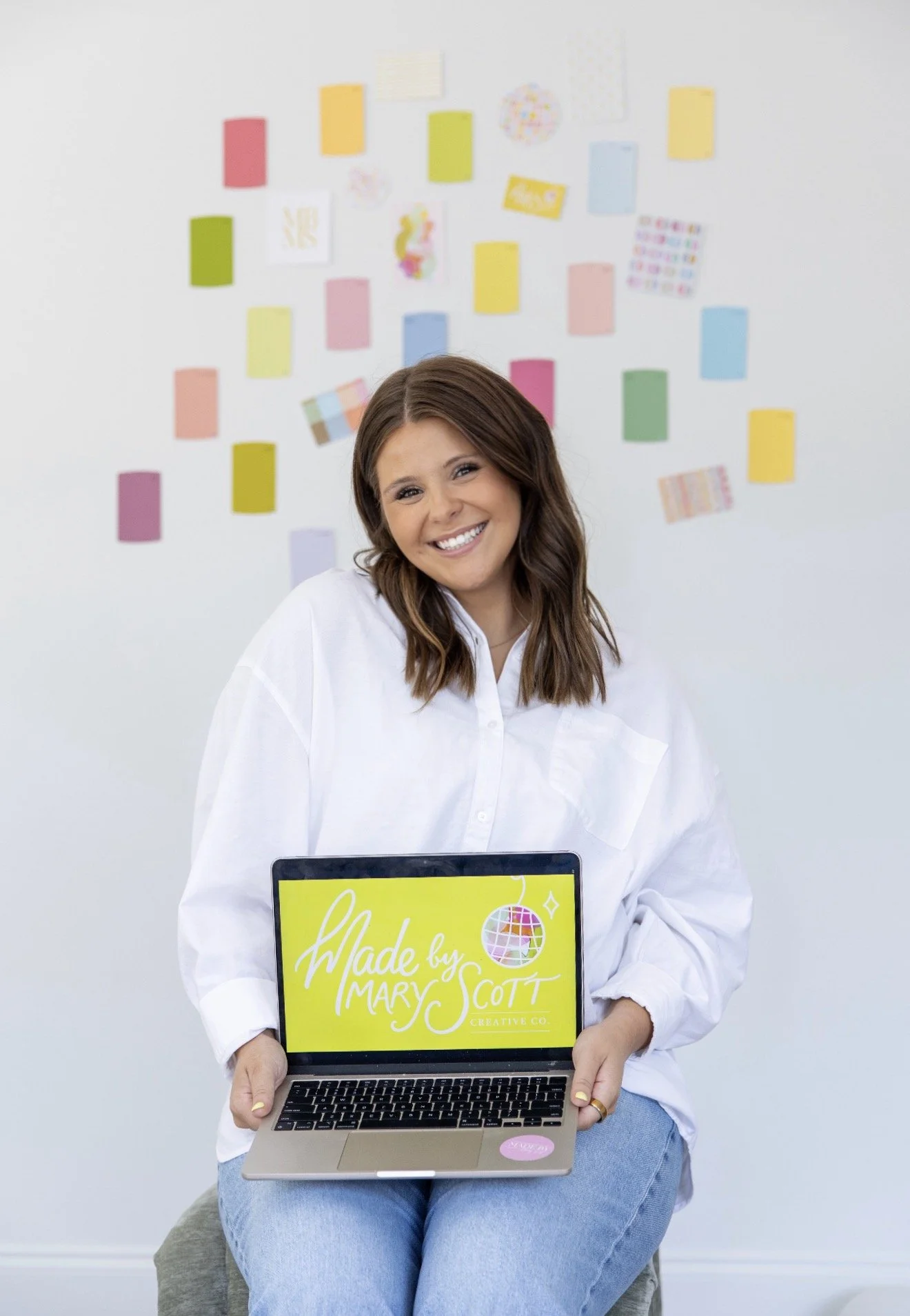 A woman with shoulder-length brown hair, wearing a white shirt and jeans, sitting on a gray cushion. She is smiling and holding a laptop with a yellow screen that reads "Made by Maryc Scott." Behind her is a light-colored wall decorated with various colorful papers and artwork.