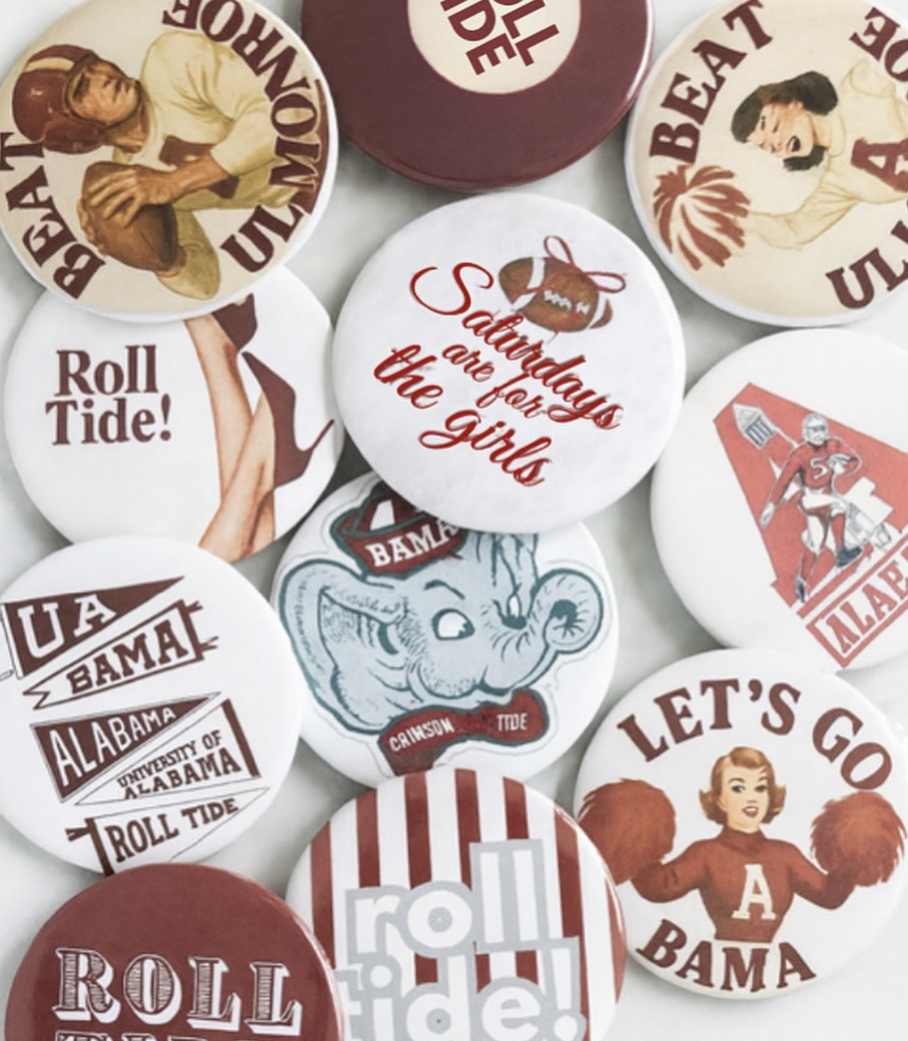 A collection of buttons and pins with various designs and slogans supporting Alabama, sports, and social themes. Designs include vintage and cartoon-style graphics, slogans like "Roll Tide!" and "Let's Go", and images of football players and other characters.