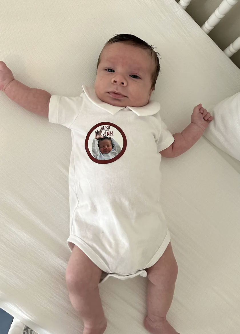 A baby lying on a bed wearing a white onesie with a sticker that has a photo of a newborn and the text "Leo says Dad" on it.