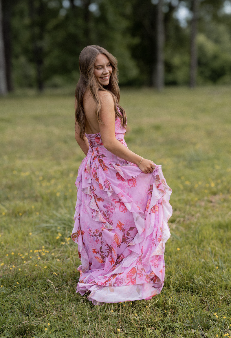 Smiling woman in a pink floral dress standing outdoors on grass with trees in the background.