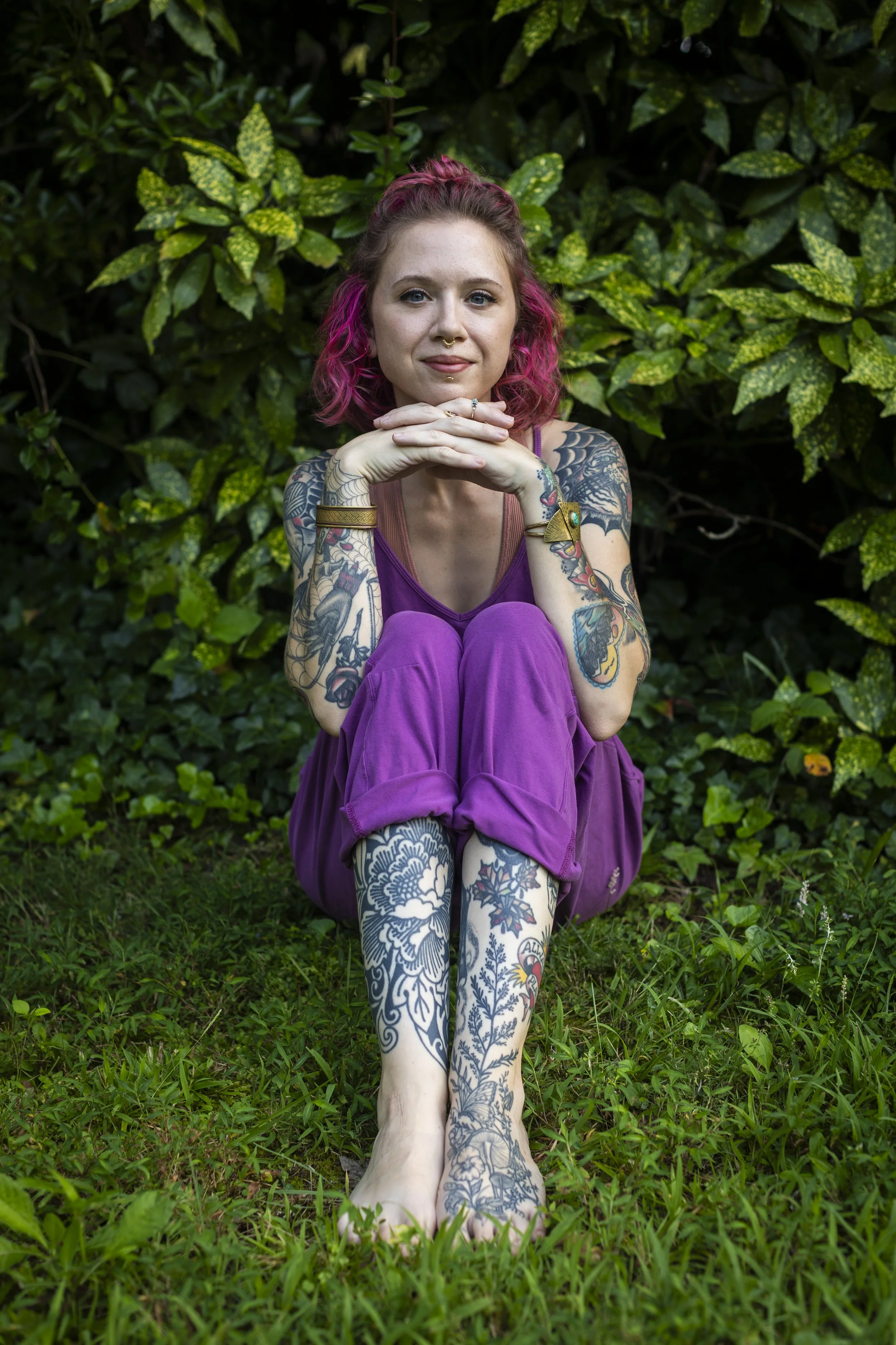 A woman with pink hair and tattoos sitting outdoors on grass with green leafy bushes in the background.