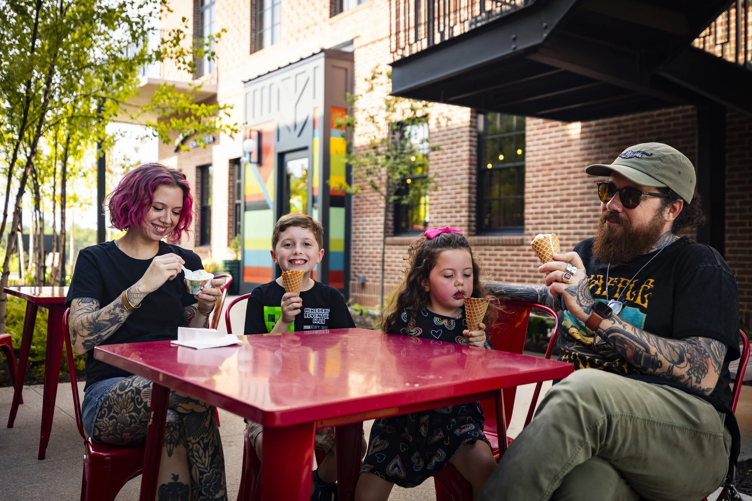 Group of five people enjoying ice cream outdoors at a red table in front of a brick building with colorful wall art and trees.