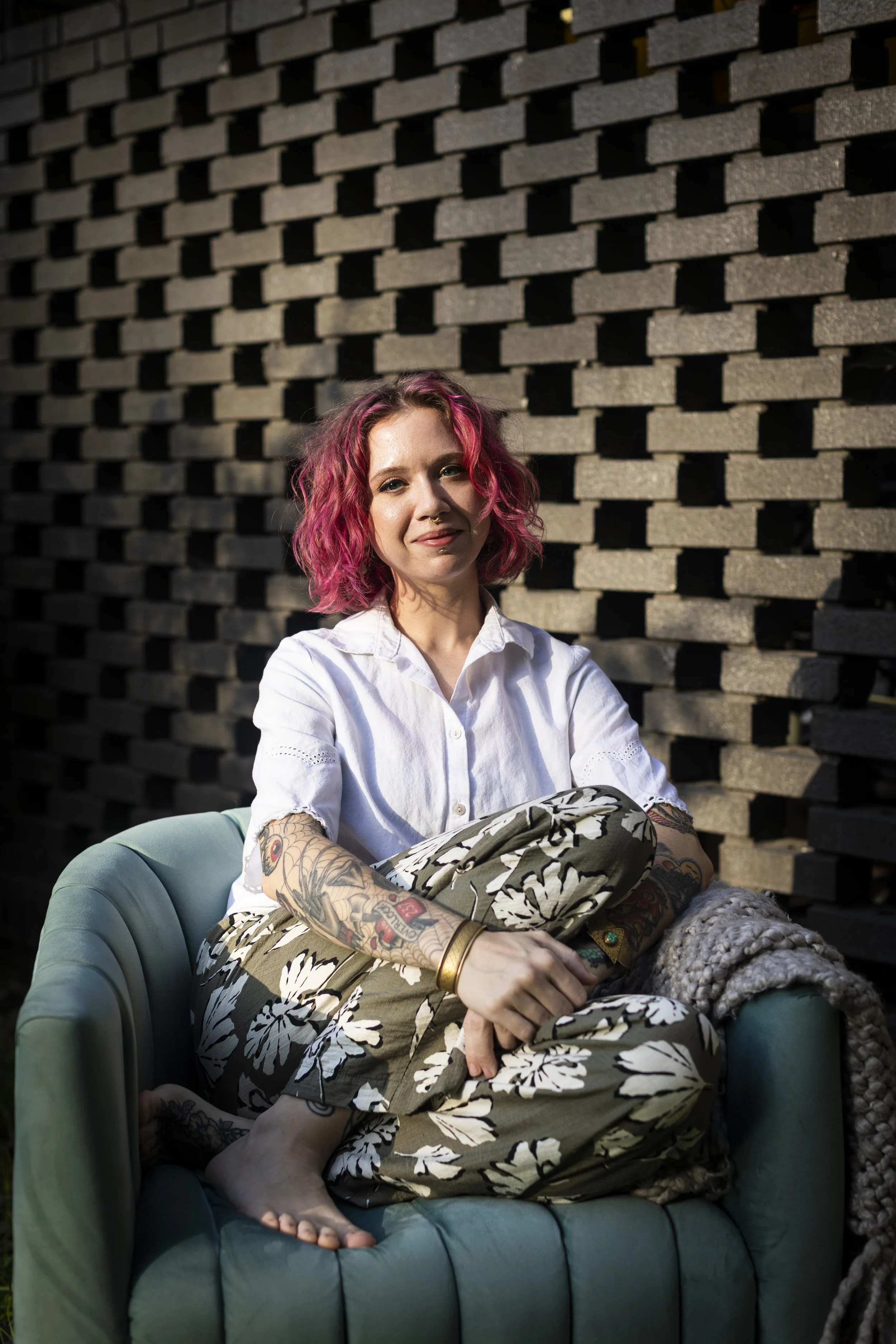 A woman with pink hair and tattoos sitting in a green armchair in front of a brick wall.