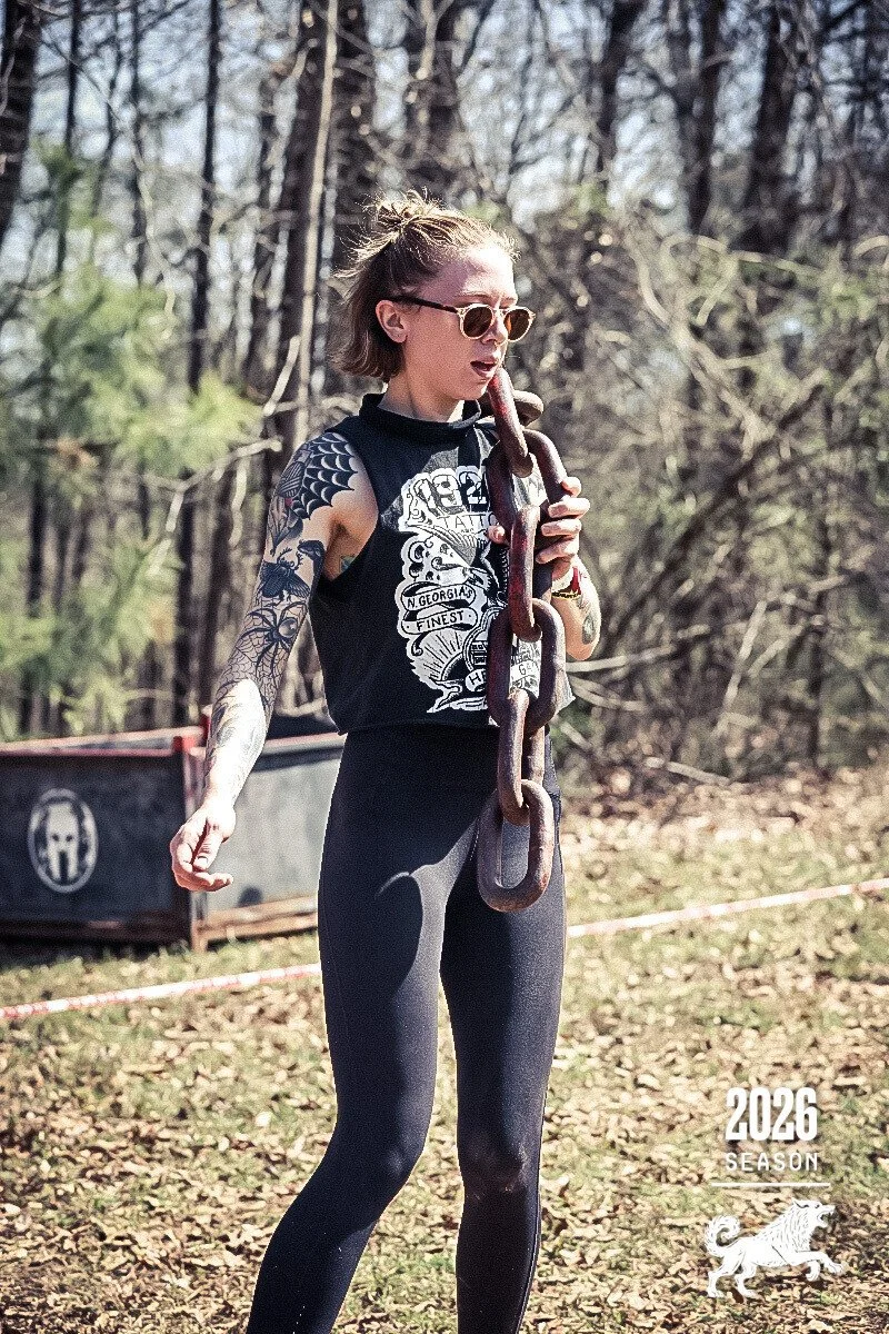 A woman with short hair, wearing sunglasses, a sleeveless black T-shirt with white designs, and black leggings, is outdoors holding a large metal chain. Trees and some equipment are visible in the background. The image includes the text "2026 SEASON" and a graphic of a lion in the bottom right corner.