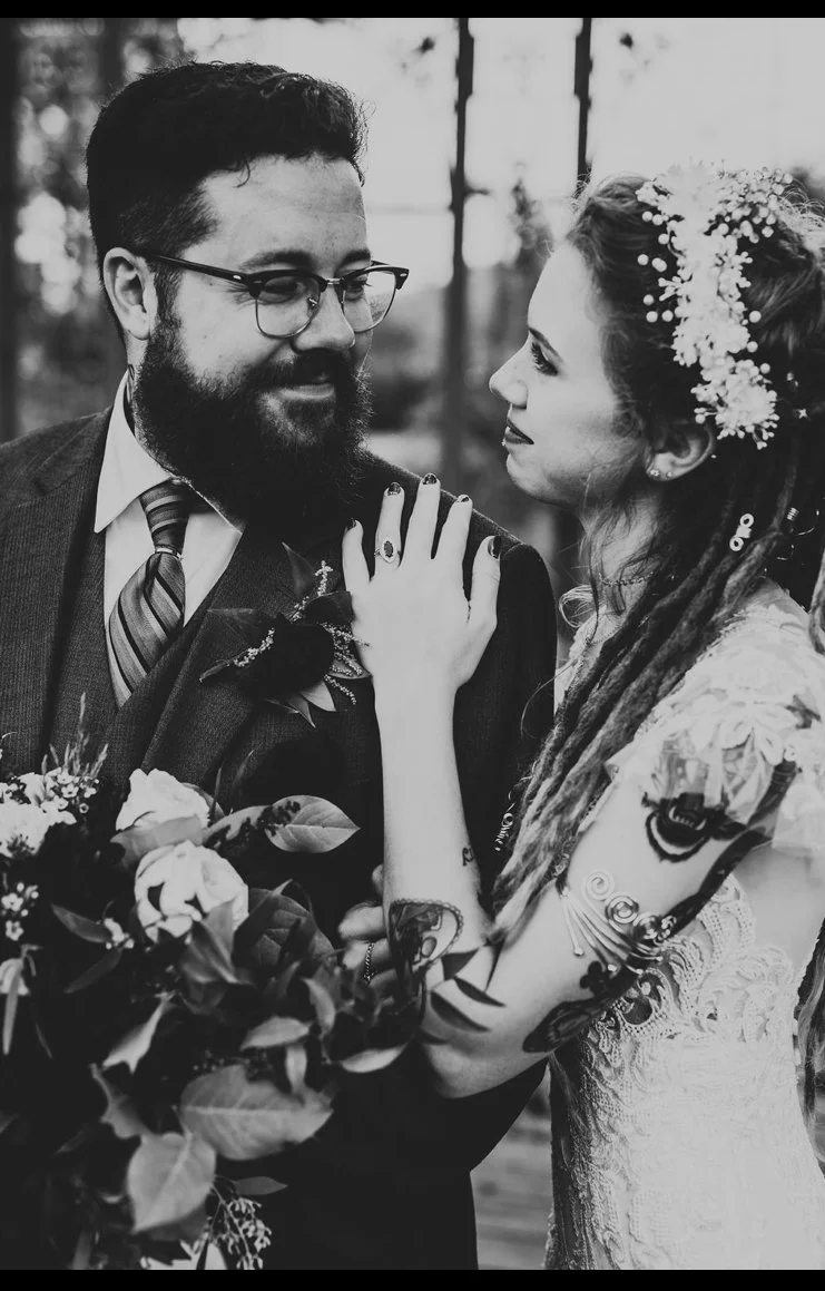 A black-and-white photo of a bride and groom looking into each other's eyes, with the bride touching the groom's shoulder and holding a bouquet, both dressed in wedding attire, outdoors with a blurred background.