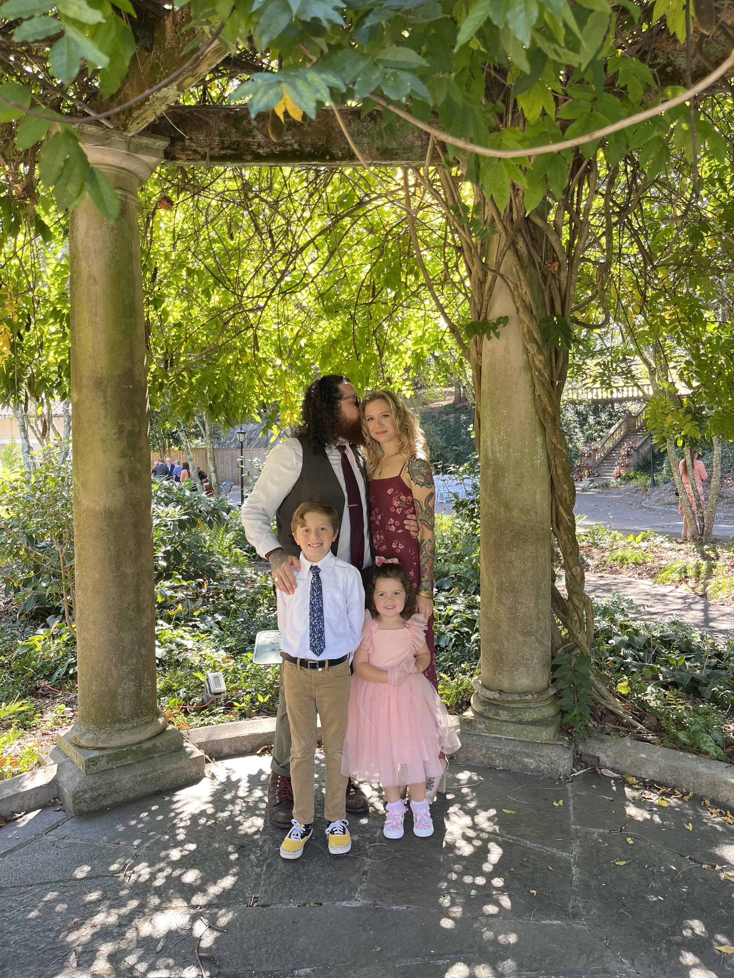 A family portrait of four standing under a green, leafy arbor with columns, in a garden setting. The adults are dressed formally, and the children are in party attire. Sunlight filters through the leaves, casting shadows on the ground.