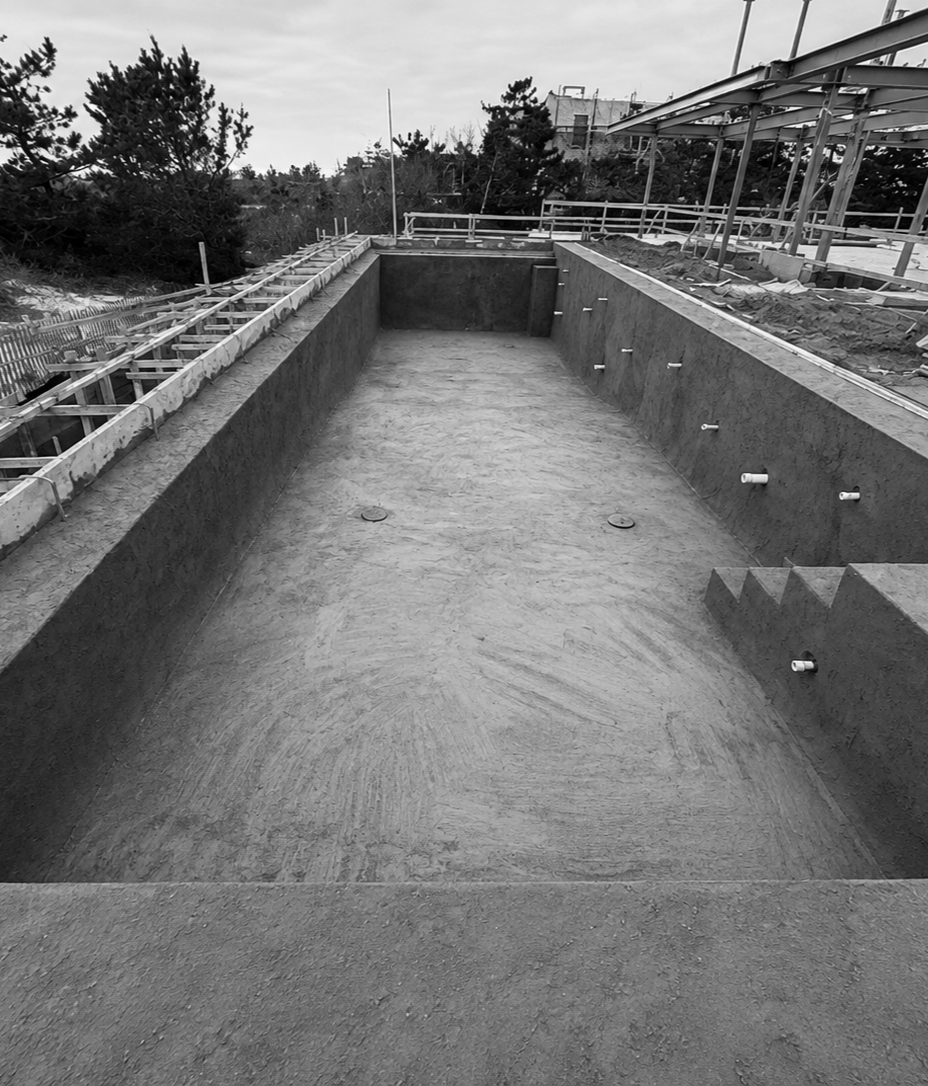 Empty swimming pool under construction, with concrete base and plumbing fixtures visible, surrounded by construction scaffolding and trees in the background.
