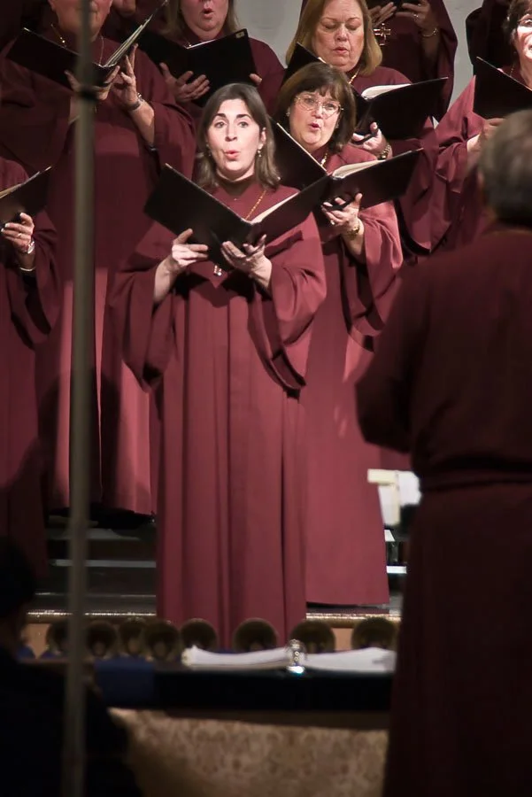 A choir of women in maroon robes singing while holding black sheet music, with a conductor in the foreground.