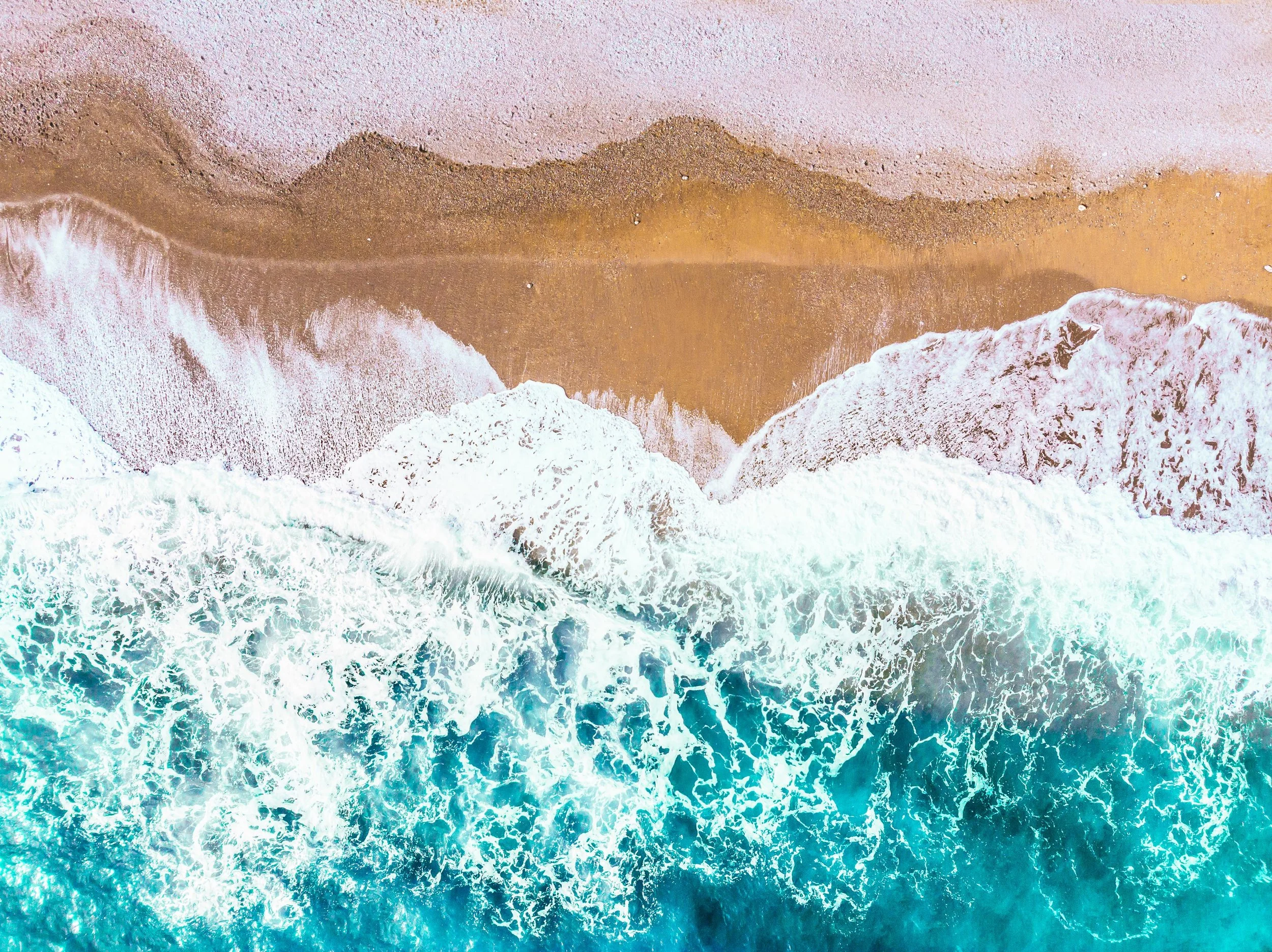 Aerial view of ocean waves crashing onto sandy beach, with white foam and clear turquoise water.