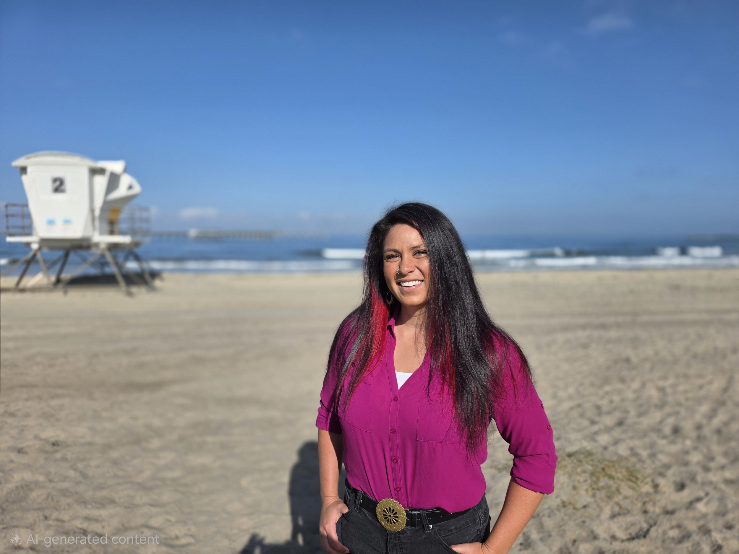 A beautiful woman with long dark hair and red streaks, wearing a pink shirt and black pants with a decorative belt, smiling on a beach with a clear blue sky, a lifeguard tower, and ocean waves in the background.