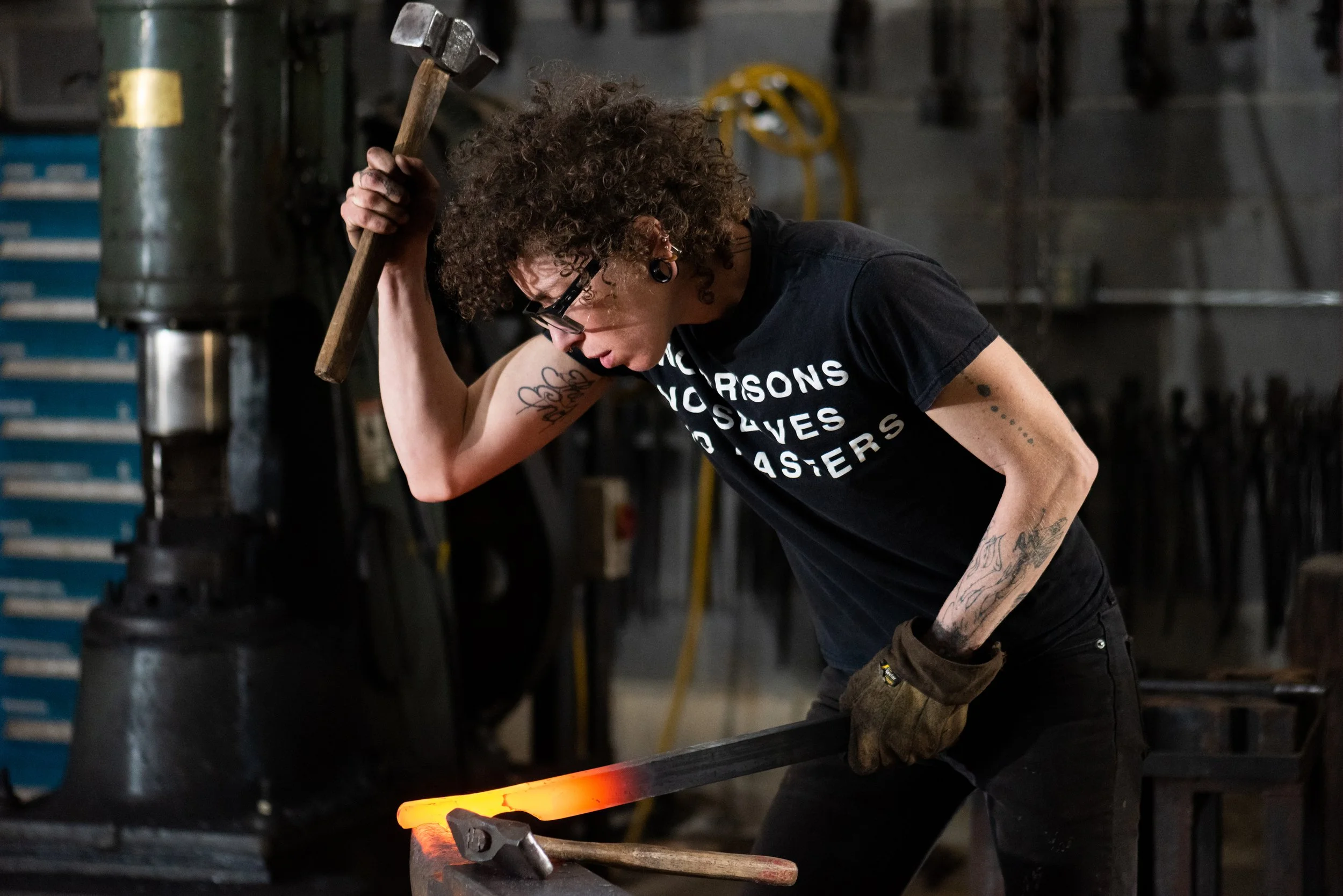 A person with curly hair, tattoos, and glasses working in a black t-shirt in a blacksmith workshop, lifting a hammer and forging hot metal on an anvil.