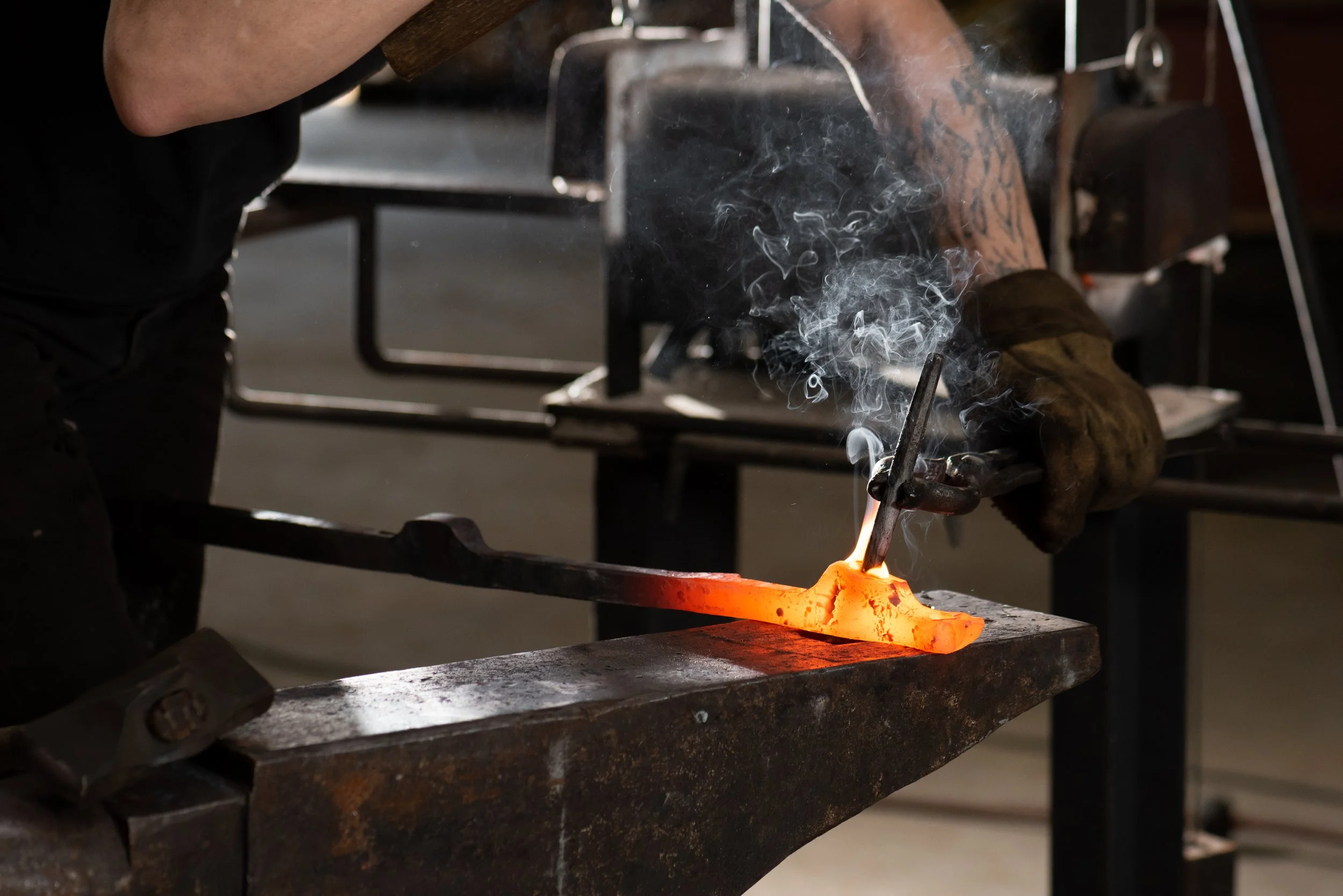 A person is forging metal using a torch and protective gloves, with smoke rising from the heated metal.