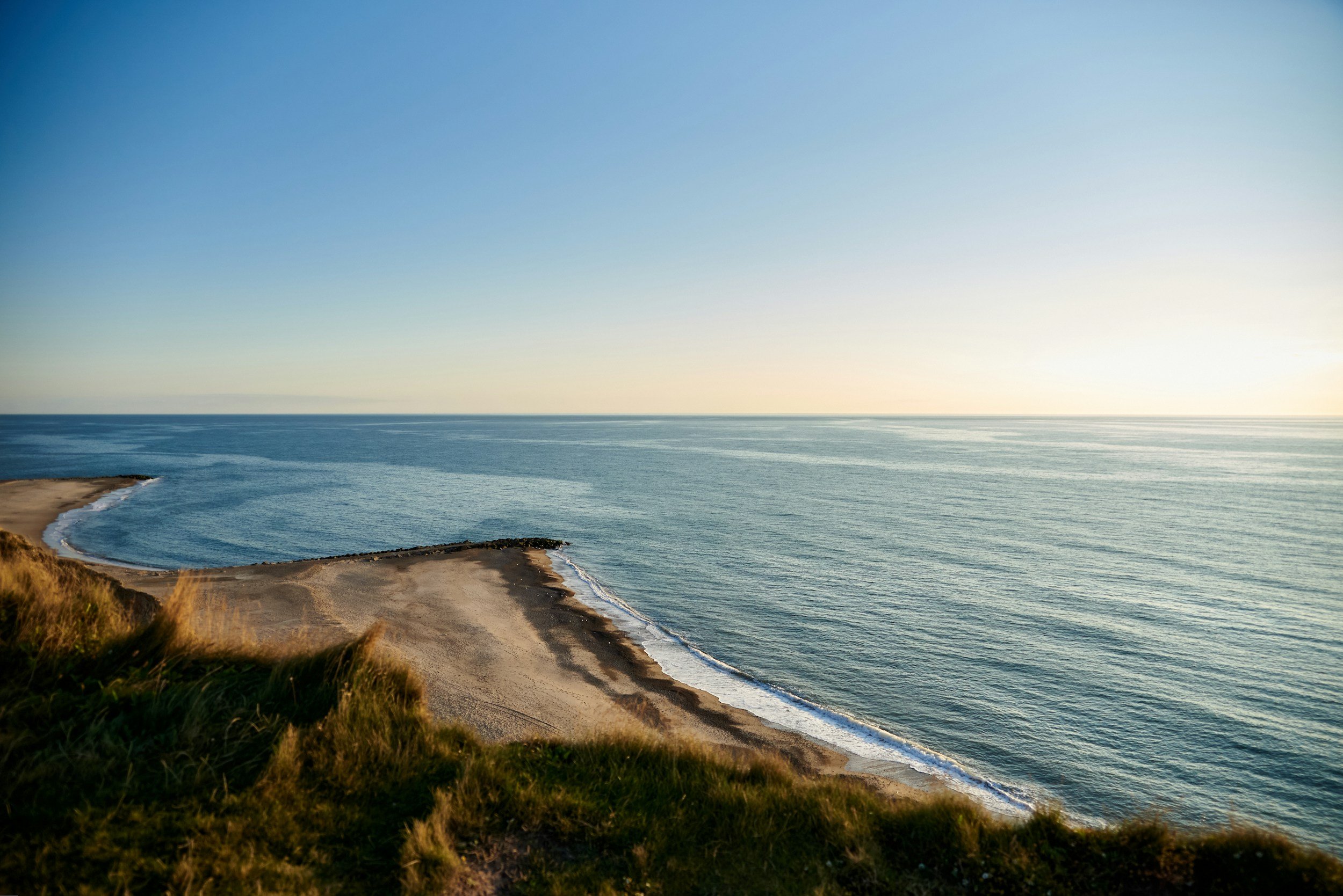 A scenic view of a coastline with a sandy beach, grassy cliffs, and calm ocean water under a clear blue sky, representing support, healing through online therapy for OCD, anxiety, and trauma.