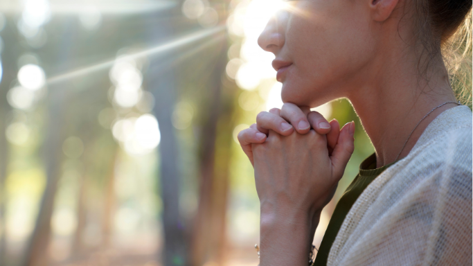 A woman standing outdoors with hands clasped in prayer, in a peaceful setting with sunlight filtering through trees, representing support, healing, and online therapy for OCD, anxiety, and trauma in Arizona.