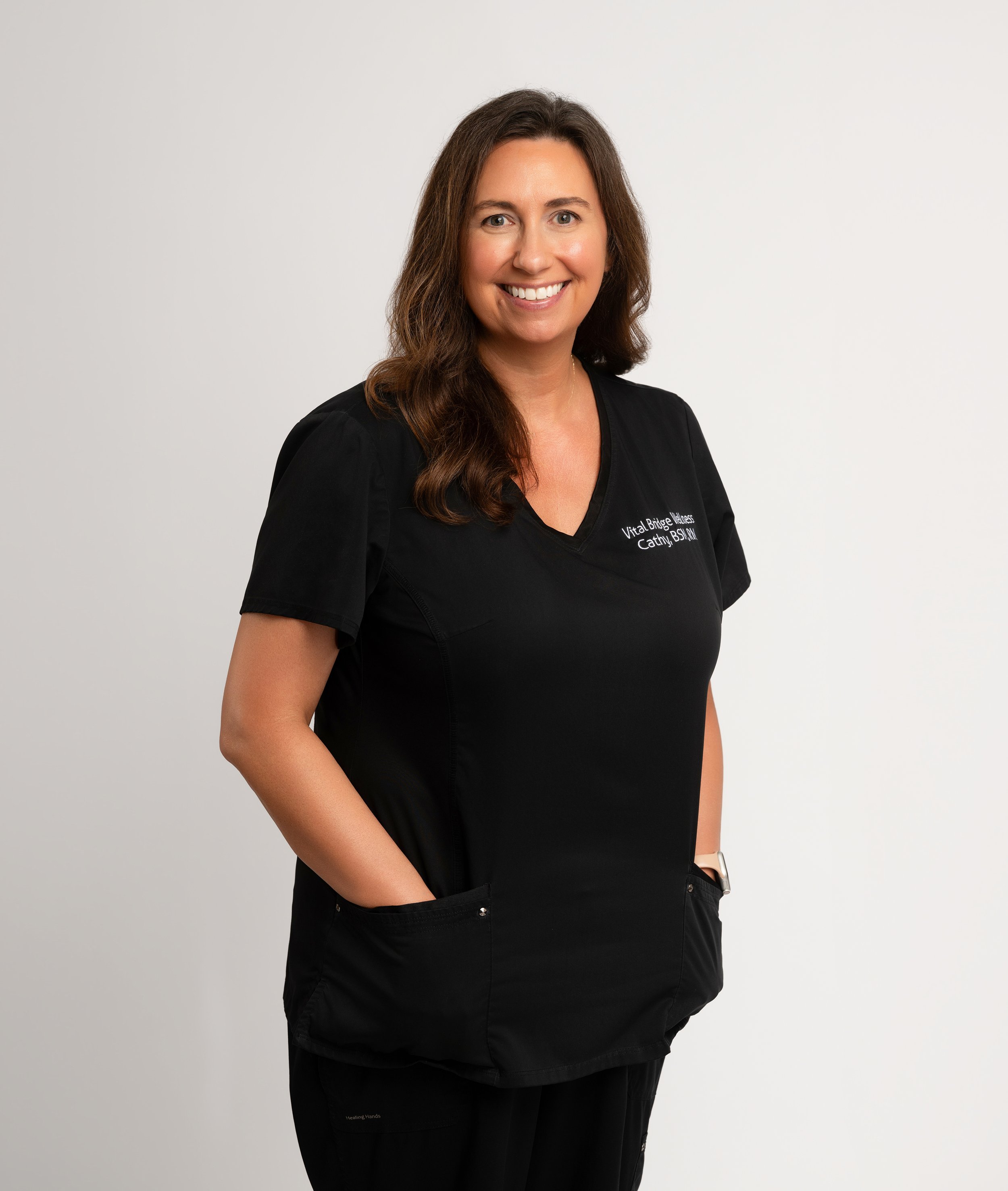 Professional woman with brown hair smiling, wearing black scrubs with embroidered text, standing against a plain white background.