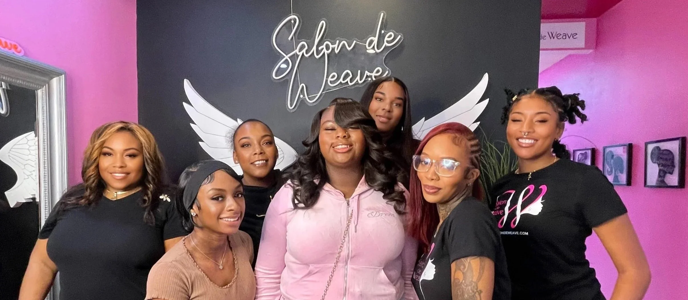 Group of seven women of diverse skin tones smiling, standing together in a pink and black salon with a "Salon de Weave" sign and wing decor in the background.