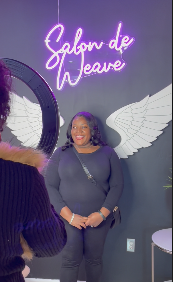 A woman smiling for a photo in front of a wall with angel wings and a neon sign that says 'Salon de Leave' in purple cursive script.