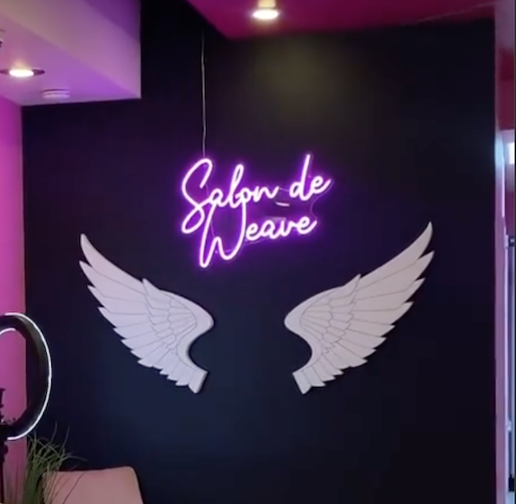 Neon sign reading 'Salon de Weave' above two white wing decorations on a dark wall.