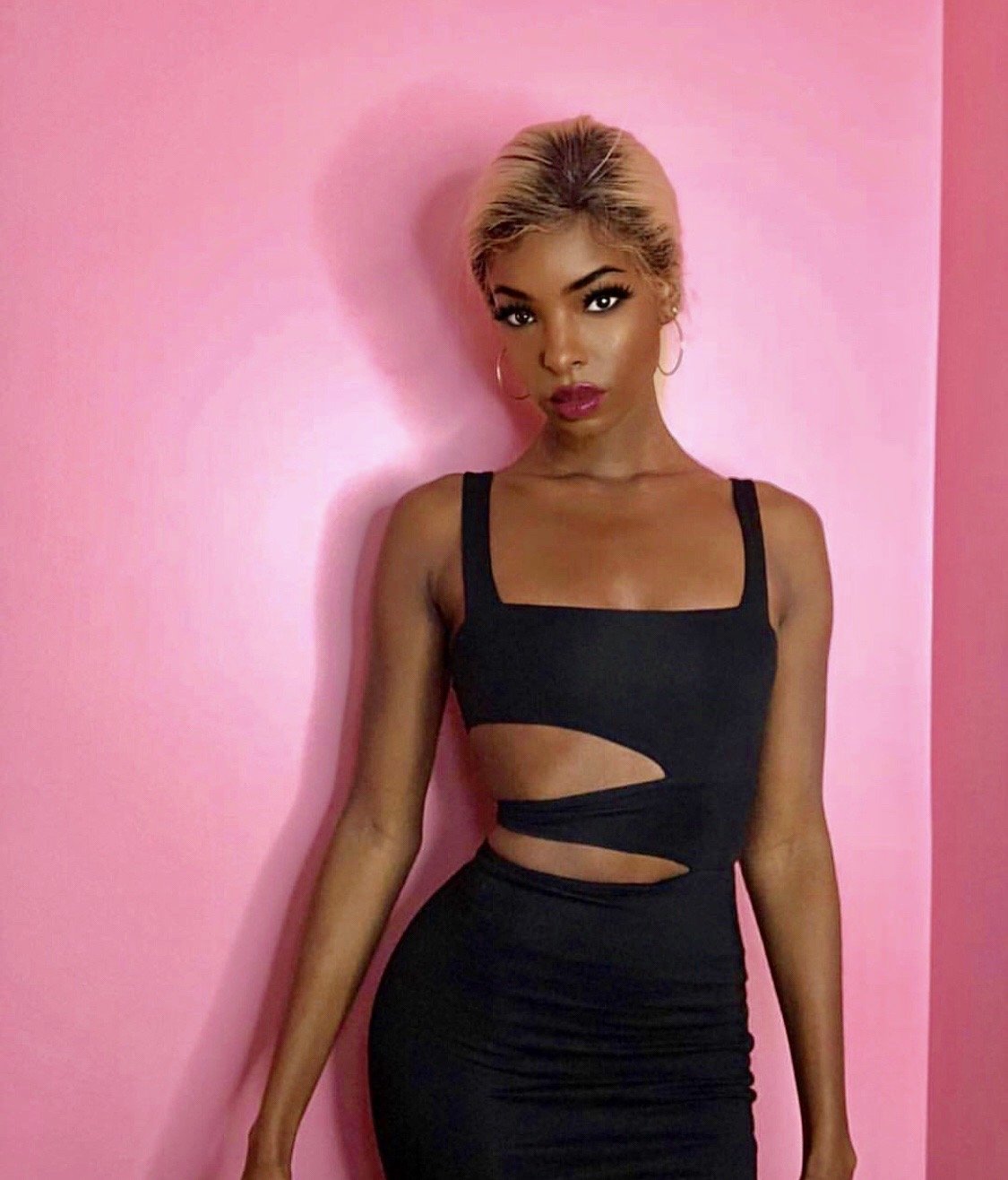 A young woman with blonde hair and hoop earrings standing against a pink wall, wearing a black dress with cutouts on the side.