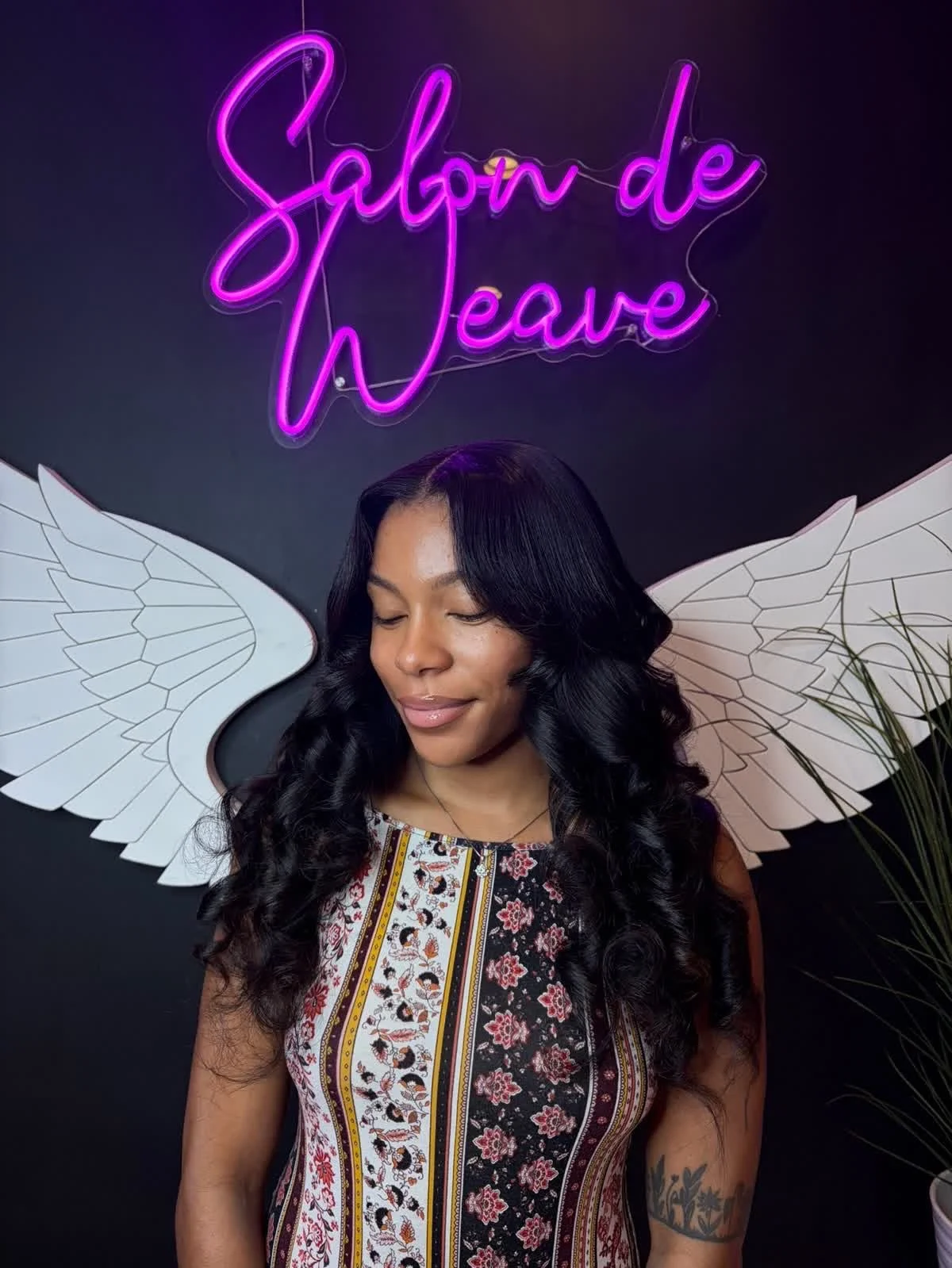 A woman with long black curly hair and a tattoo on her arm, with white angel wings behind her, stands in front of a black wall. She has her eyes closed and a gentle smile. Above her, a neon pink sign reads 'Salon de Wowe'.