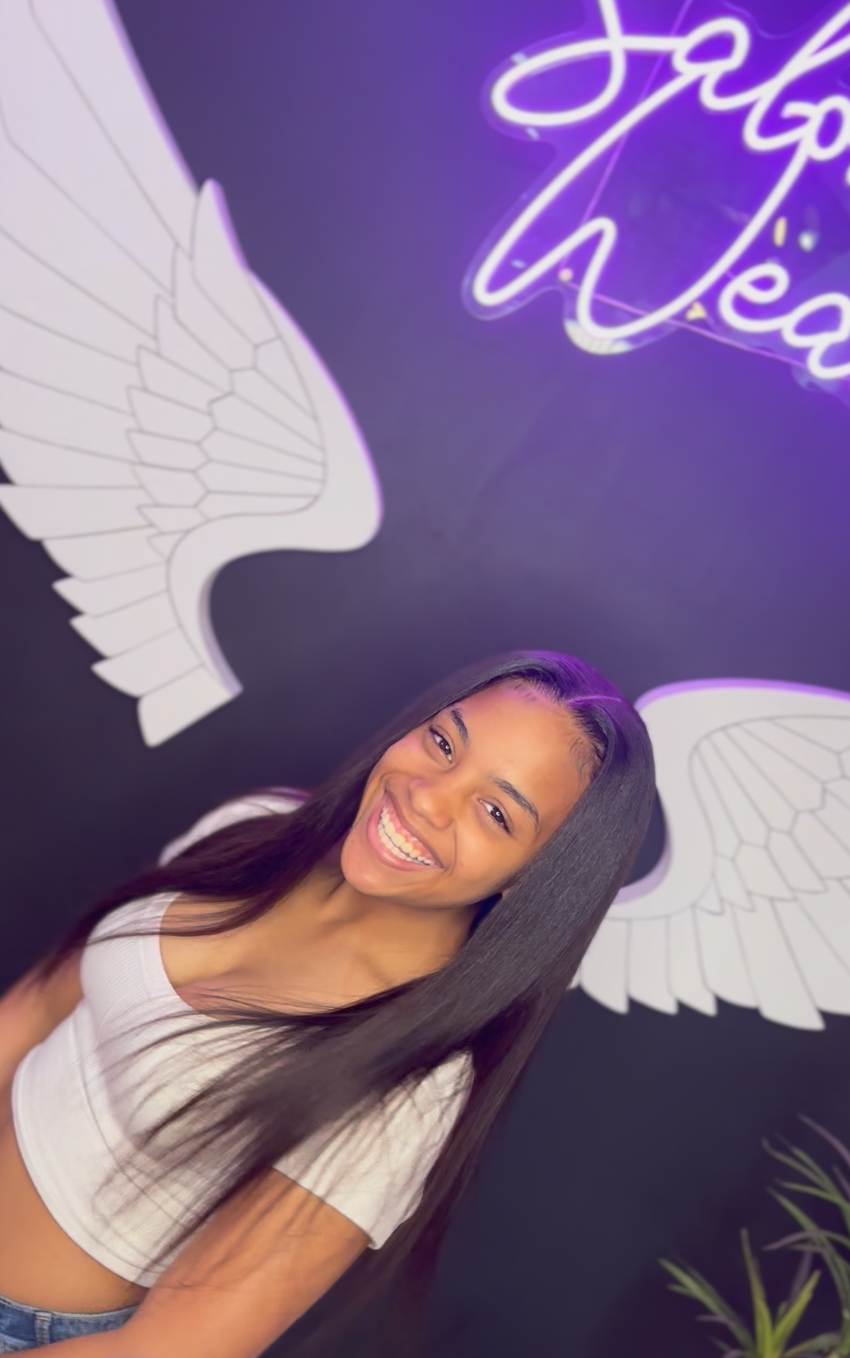 A woman with long straight dark hair smiling in front of a black wall decorated with neon angel wings and a neon sign.